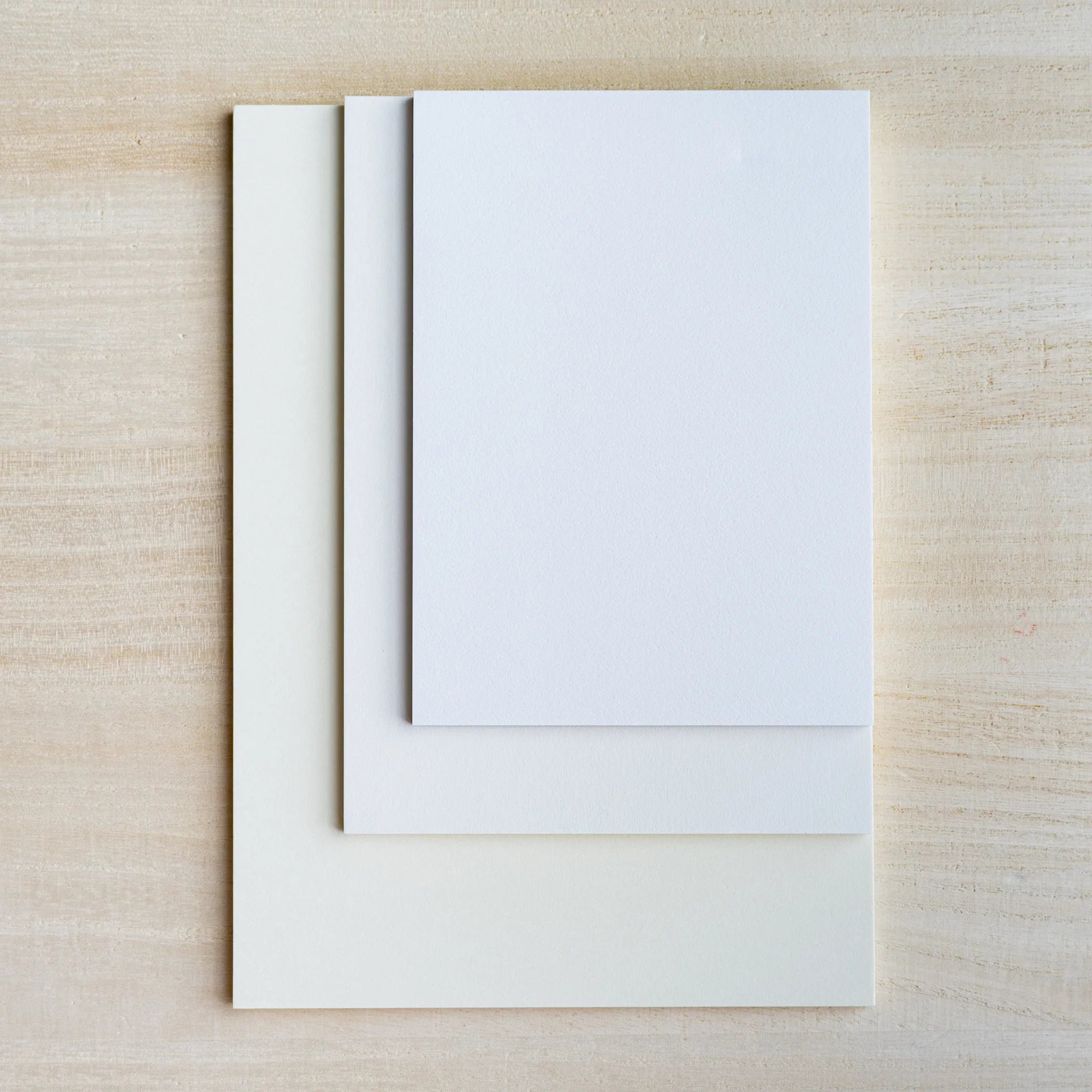 Three blank sample sheets in cream, gray and white stacked at different sizes on light wood