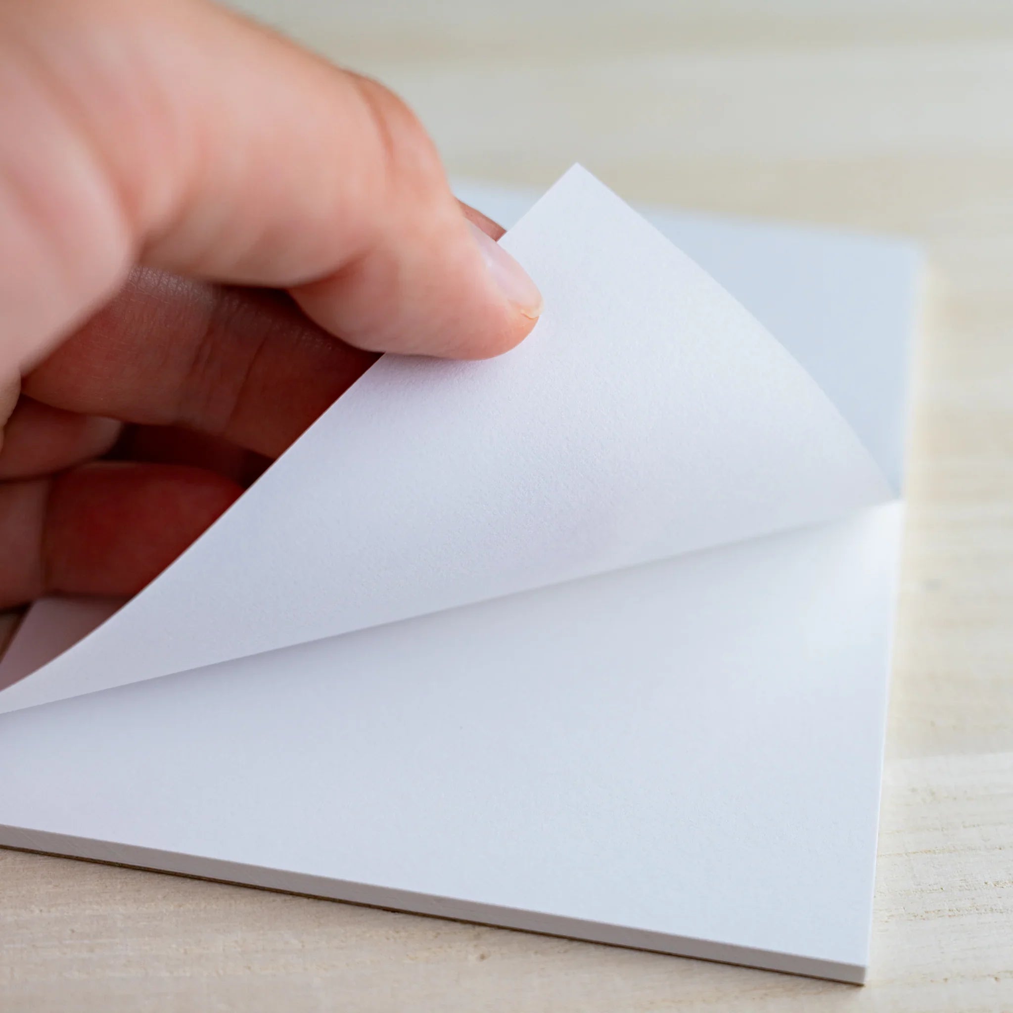 Fingers peeling back a white sheet from the bound writing pad, light wood background