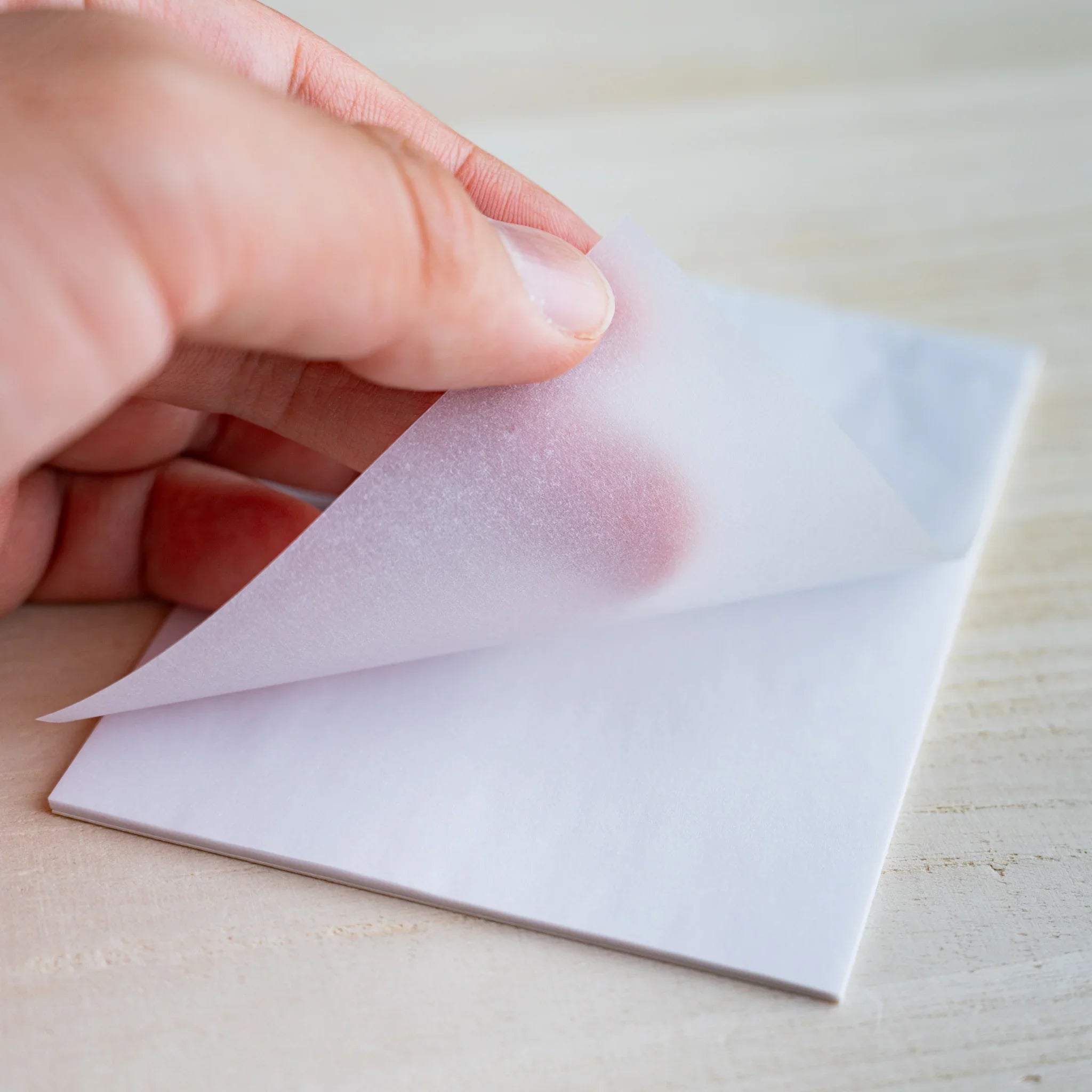 Hand lifting a translucent sheet, fingers showing through the semi-transparent paper, on wood