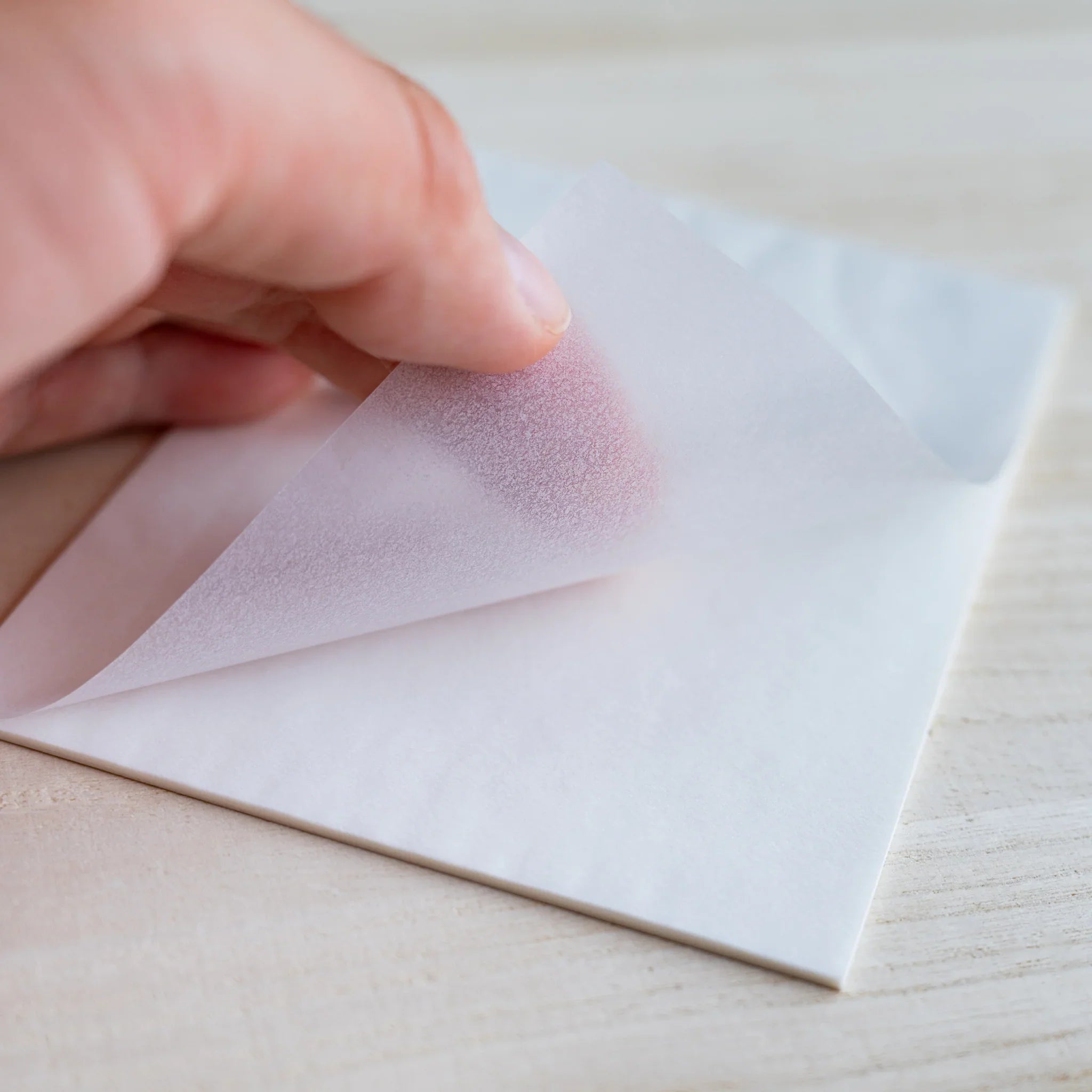 Fingers peeling a translucent sheet, hand visible through the semi-transparent paper, on wood