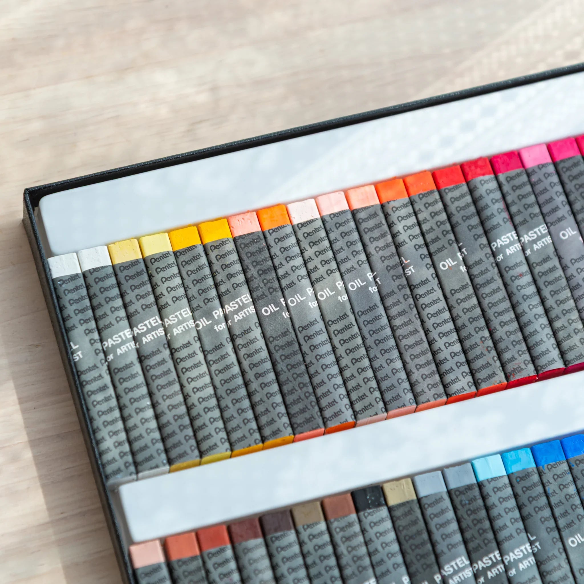 Angled view of open Pentel oil pastel box showing warm color tips in top row: white, yellow, orange, red and pink