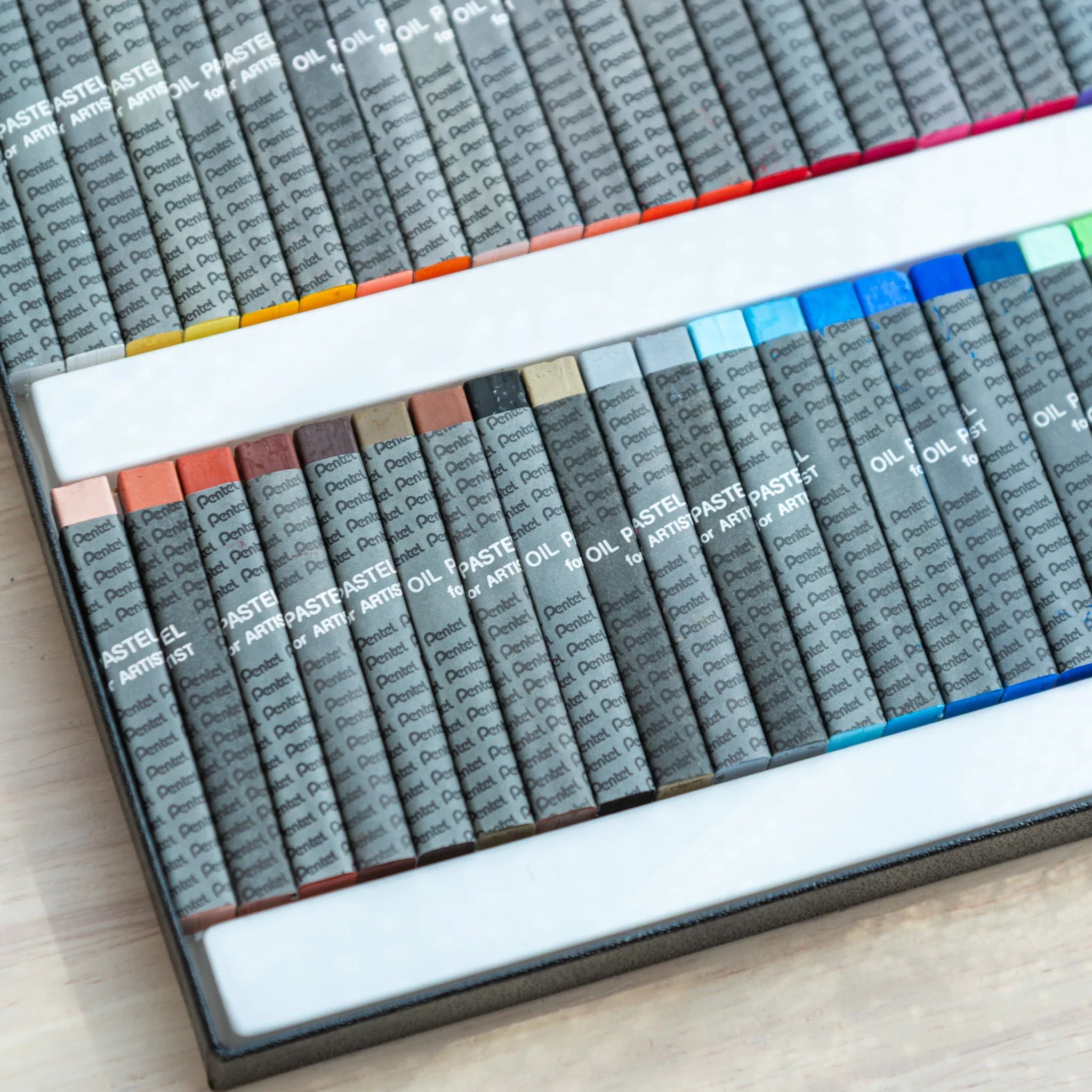 Angled close-up of Pentel oil pastel box showing earth-toned colors in lower tray including browns, grays, and blue tones
