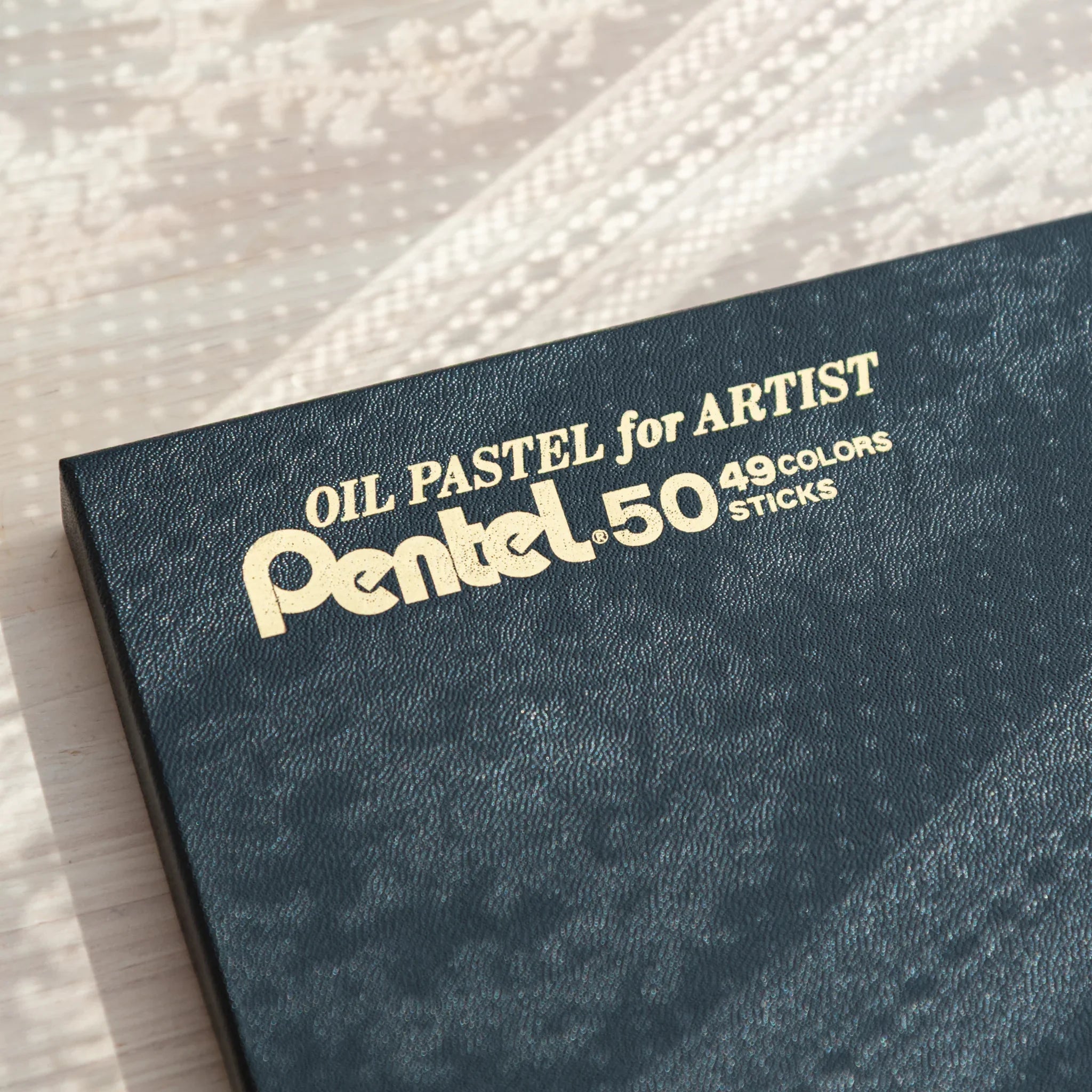 Angled close-up of Pentel Oil Pastel for Artist box lid corner showing gold foil OIL PASTEL FOR ARTIST text and teal texture