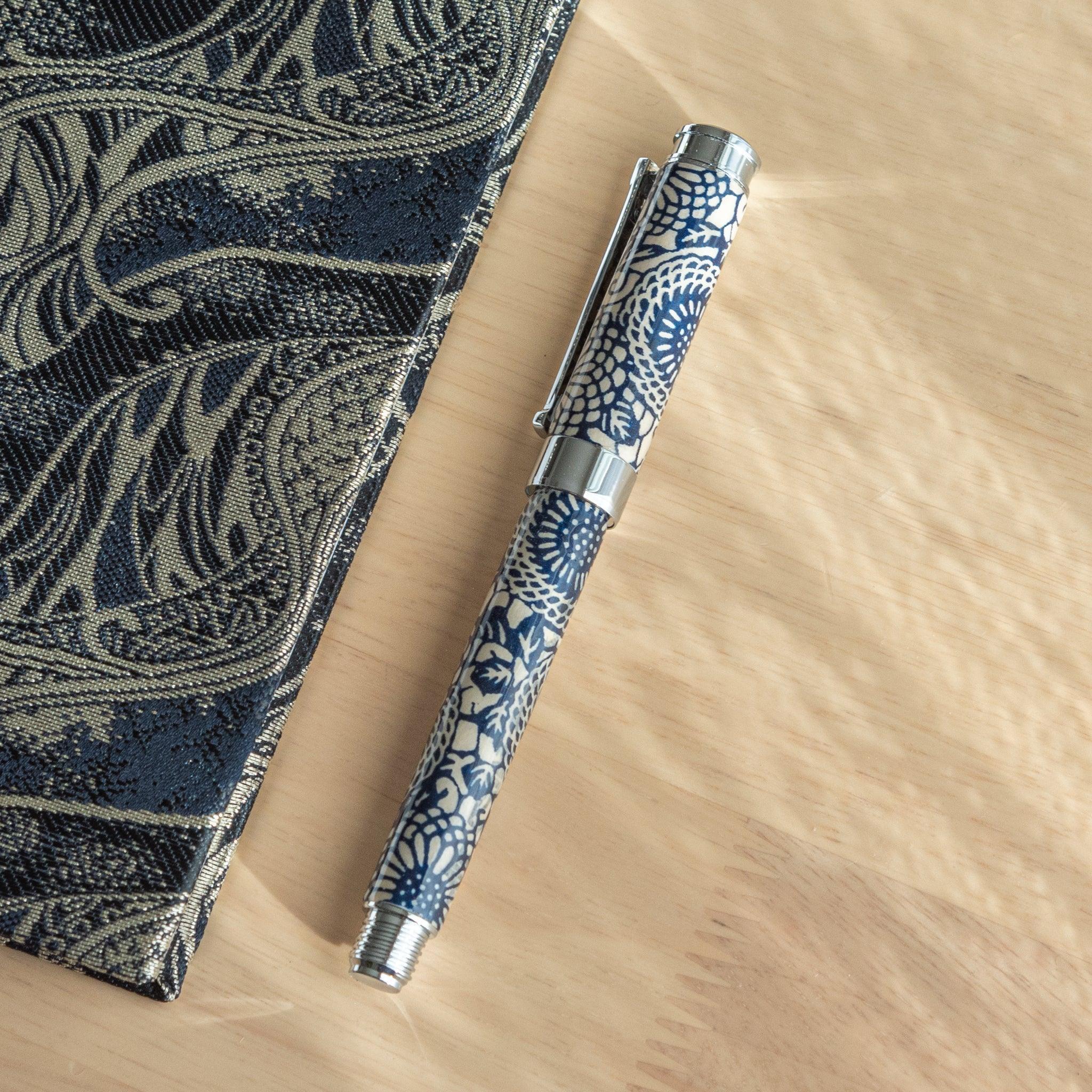 A Japanese stationery fountain pen with intricate blue floral designs, resting diagonally on a wooden surface near a notebook.