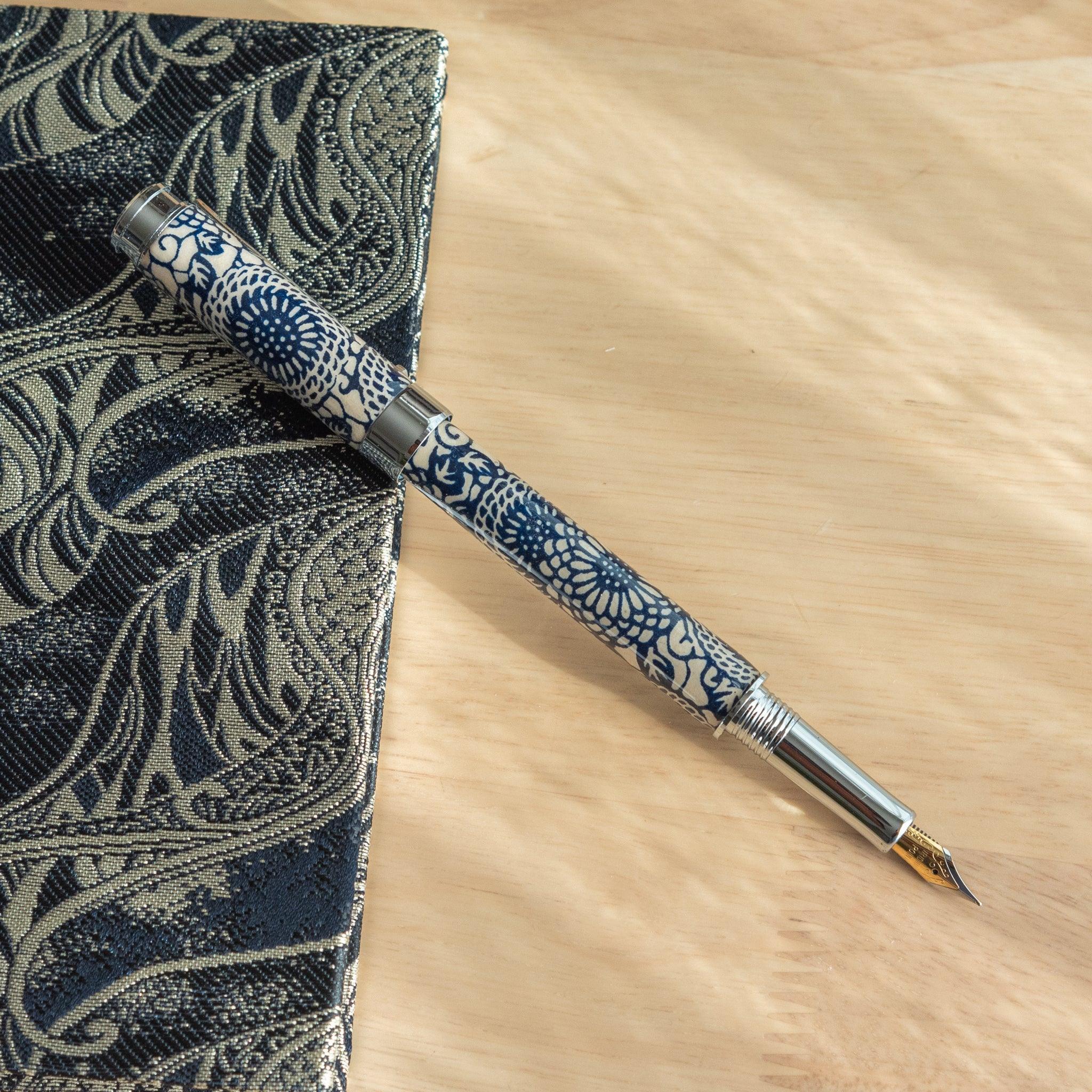 A Japanese stationery fountain pen with blue floral patterns and a silver nib, placed uncapped on a wooden surface beside a notebook.