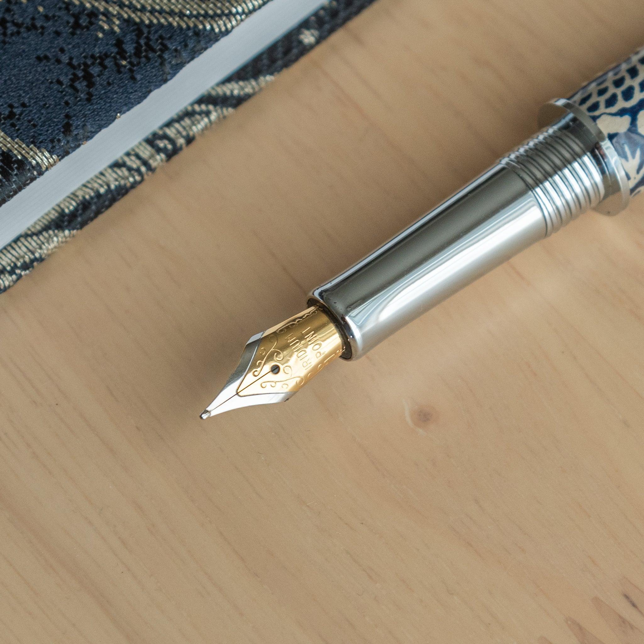 Close-up of the gold nib of a Japanese stationery fountain pen with silver trim, placed on a wooden surface next to a notebook.