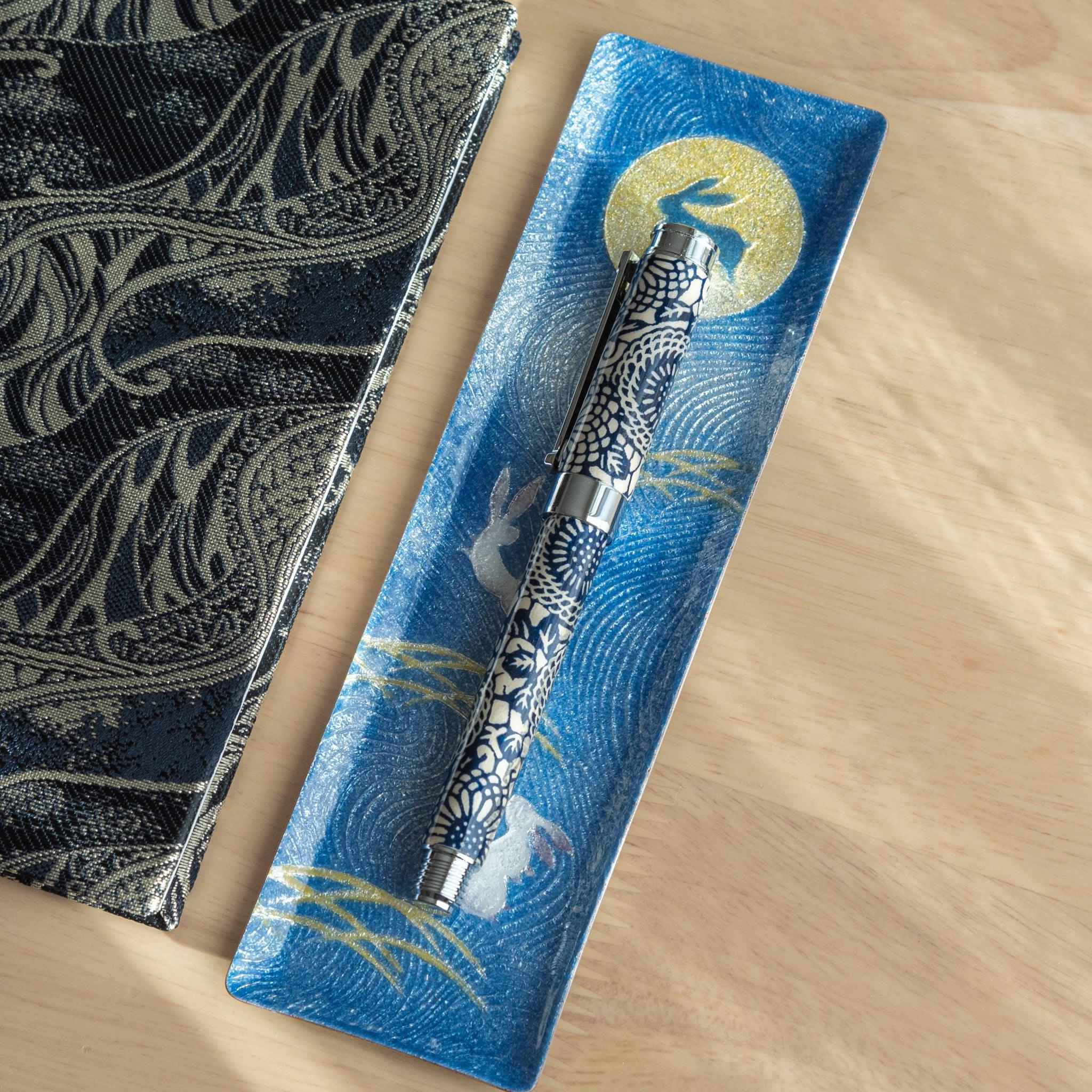 A Japanese stationery fountain pen with blue chrysanthemum patterns, resting on a blue tray with moon and rabbit designs.