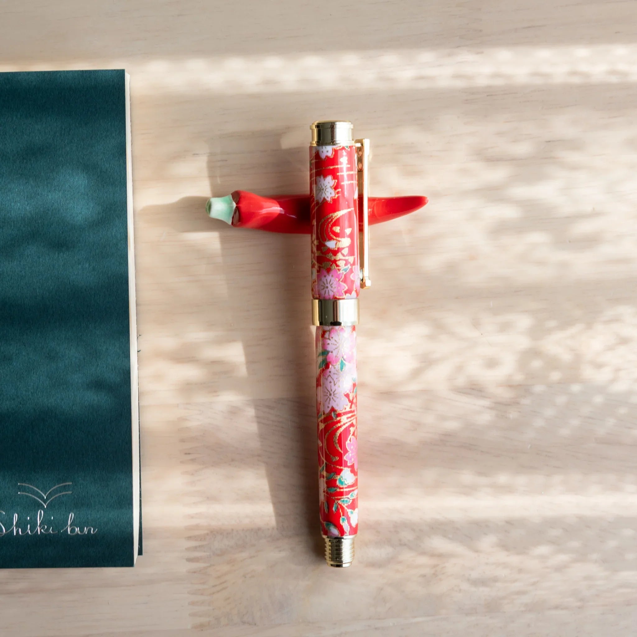 Red pepper-shaped ceramic pen rest with intricate floral pattern and mint green stem, horizontal writing holder