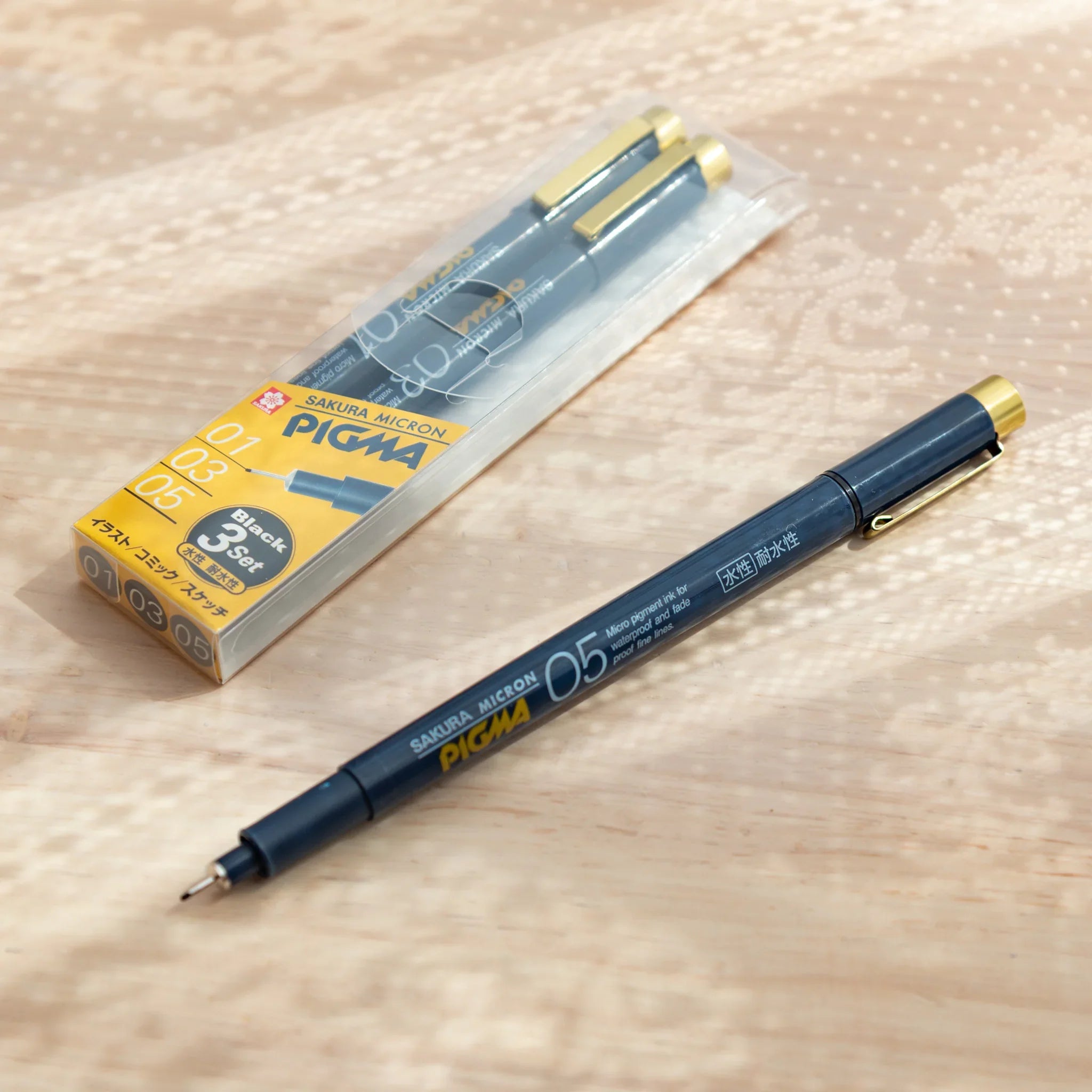 Pigma Micron fineliner pen set packaging with one pen removed showing dark grey barrel, fine tip, and gold cap