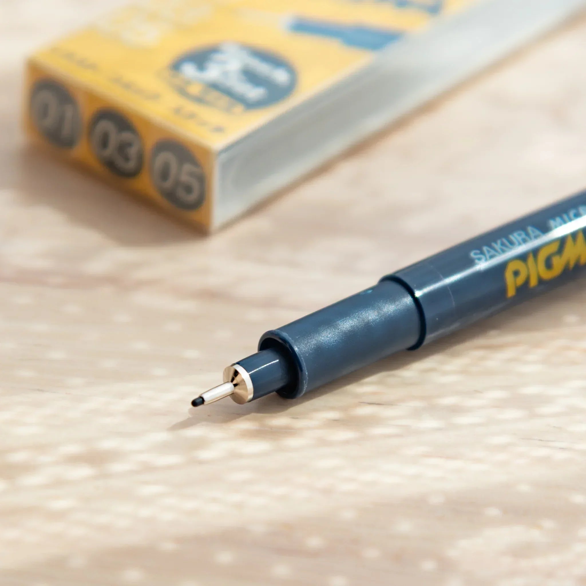 pigma micron pen close-up with fineliner nib extended on grey barrel with yellow logo, blurred packaging behind