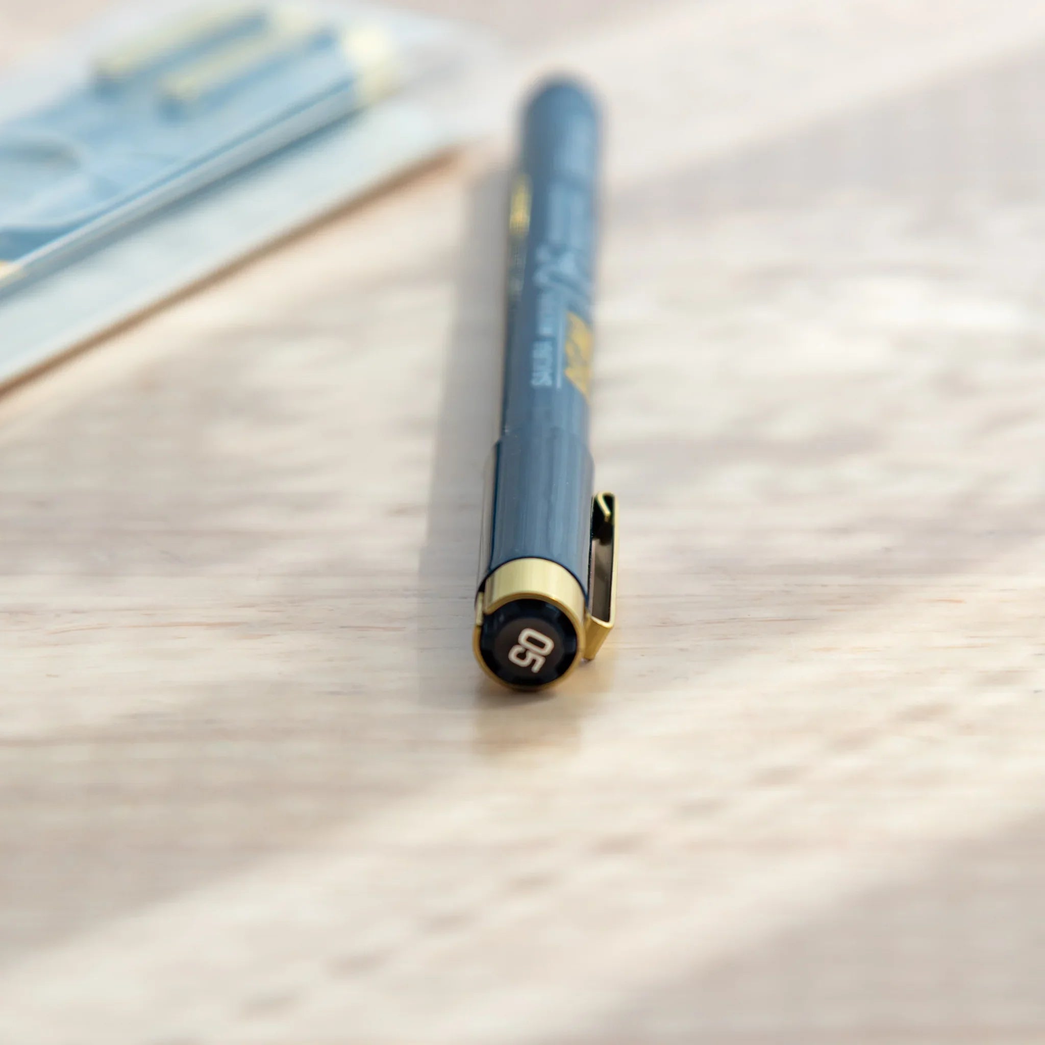 Pigma Micron fineliner pen rear view with 05 label on cap, dark grey barrel, gold clip and blurred packaging