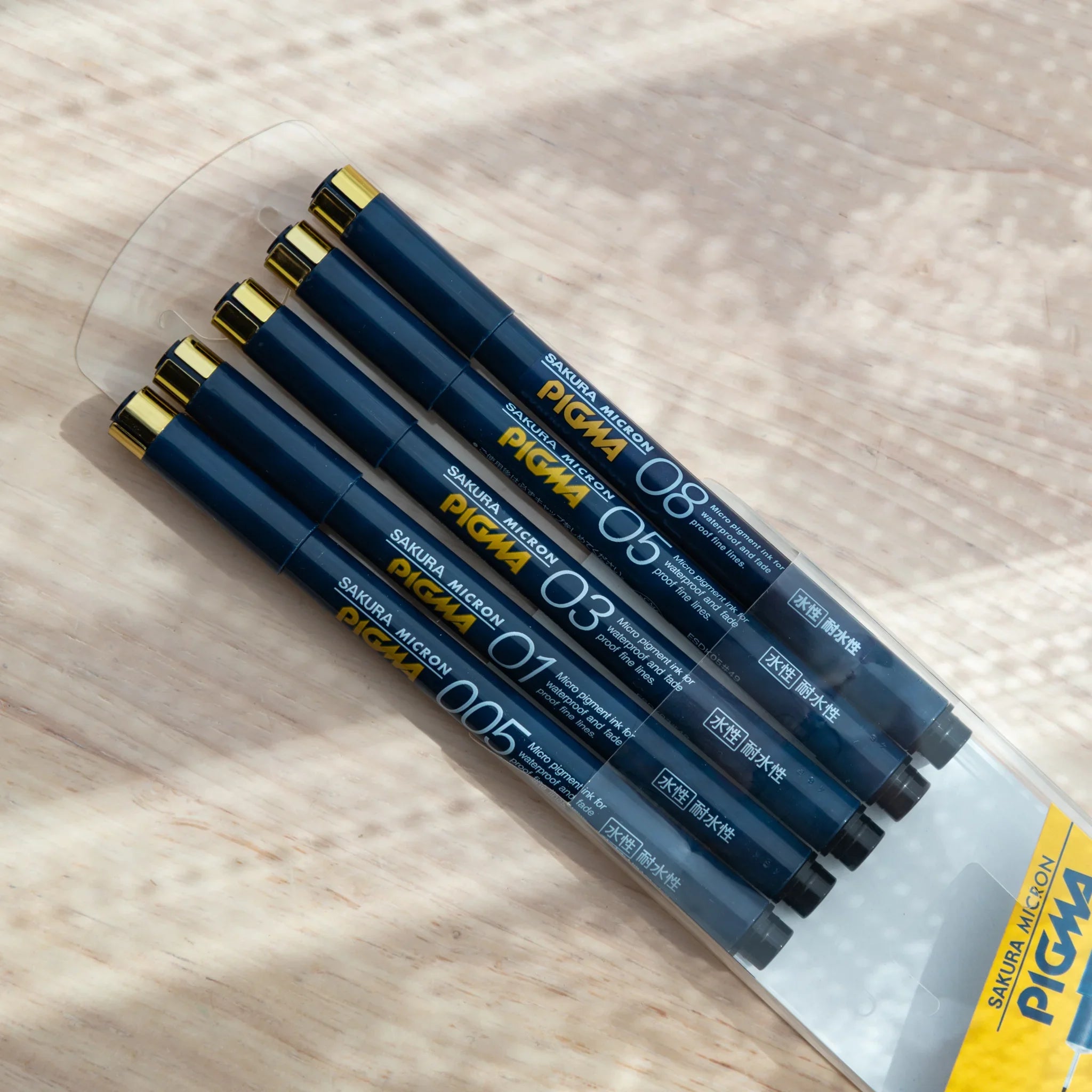 Five Sakura Pigma Micron fineliner pens fanned out of their sleeve showing dark navy barrels and gold clip caps
