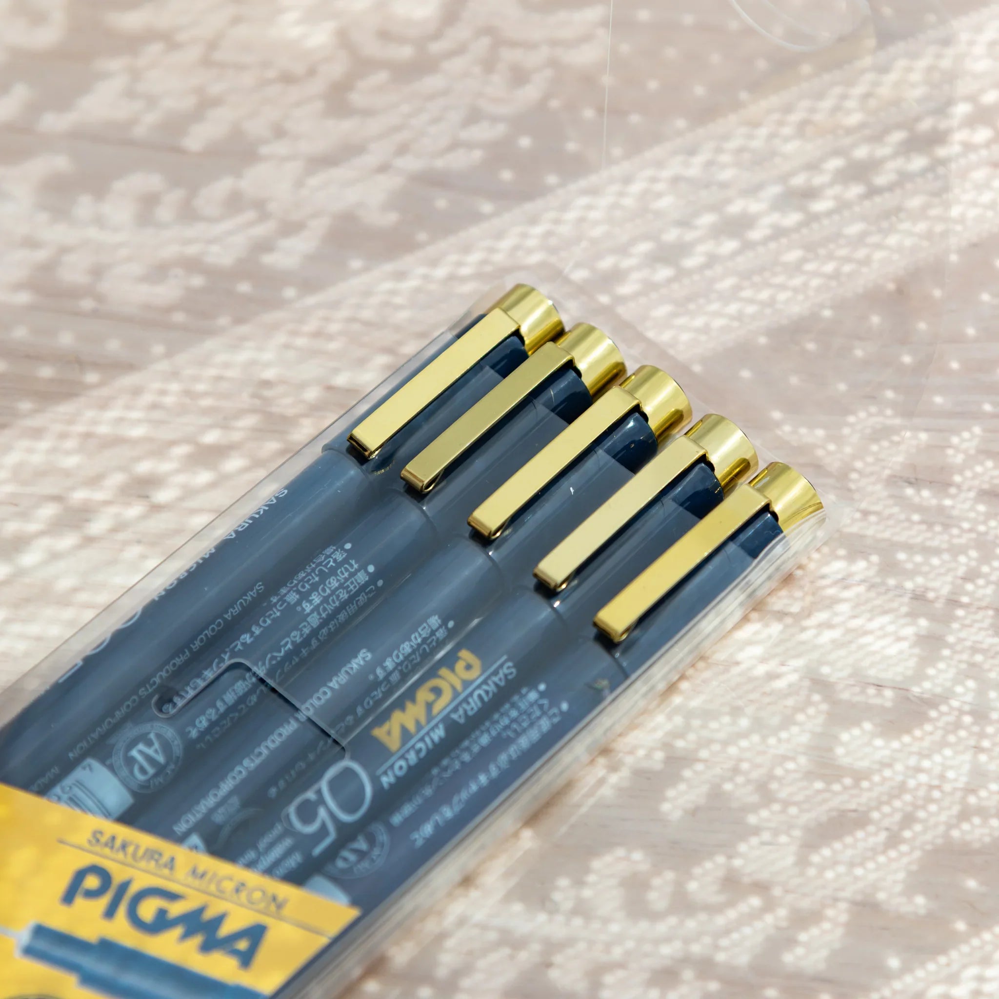 Close-up of five Pigma Micron pen cap ends with gold clips inside the clear plastic sleeve, arranged in stepped formation