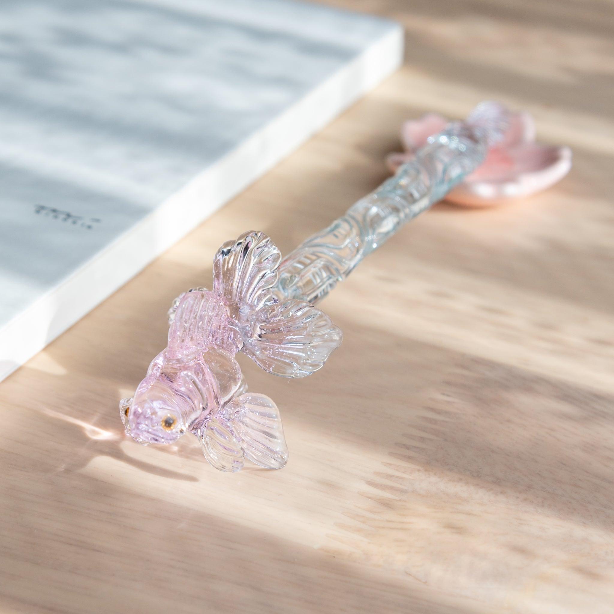 A Japanese glass dip pen with a pink goldfish motif and intricate glass details rests on a pink maple leaf-shaped holder beside an MD notebook on a wooden desk.