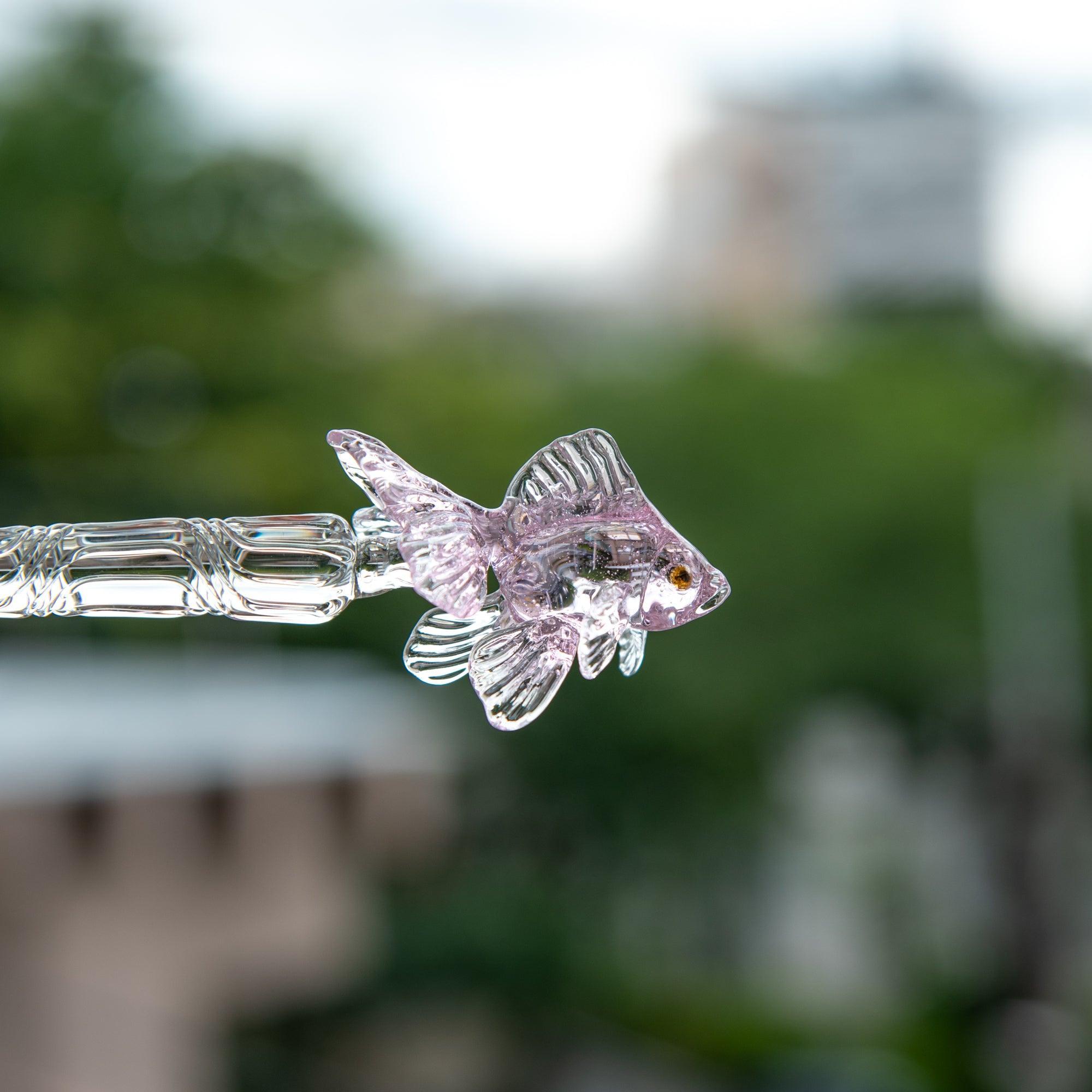 The pink goldfish glass pen resting on an open notebook, its translucent body and fish design casting a soft shadow across the page.