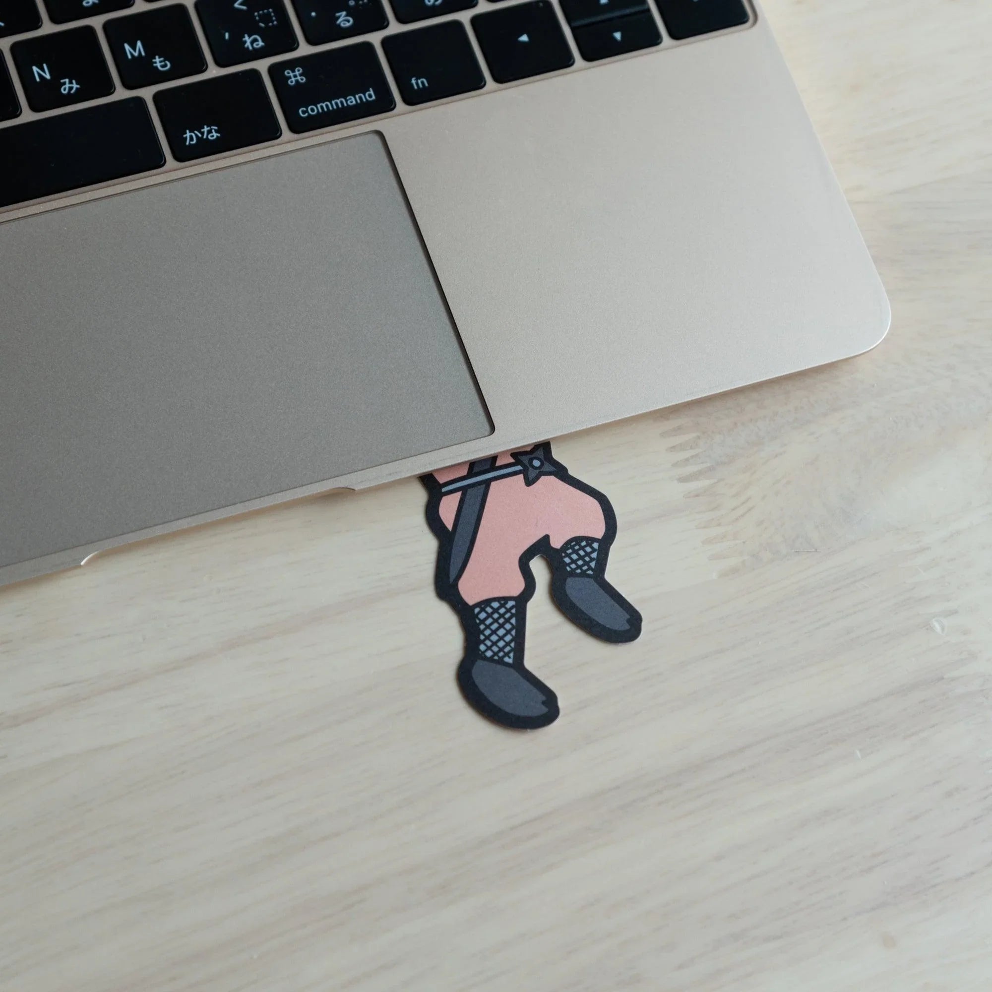 Pink ninja die-cut message card tucked under a gold MacBook laptop with only the lower body and boots visible, on a wood desk