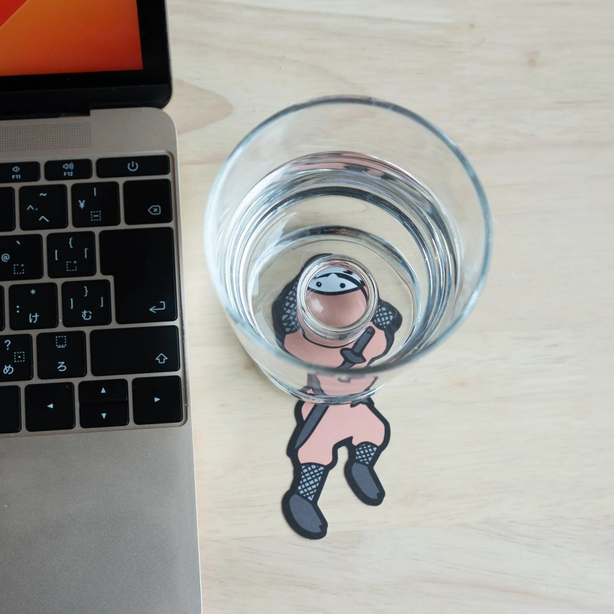 Pink ninja die-cut card under a glass of water on a desk, the ninja head magnified and visible through the glass base
