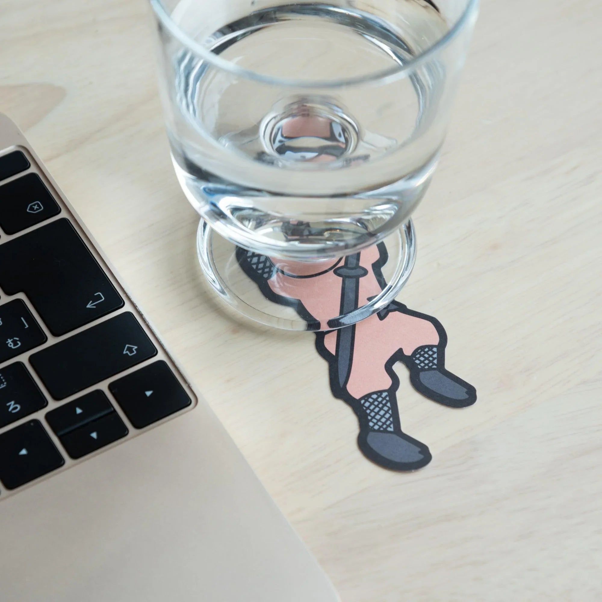 Pink ninja die-cut card under a clear glass of water beside a laptop, lower body of ninja visible outside the glass base