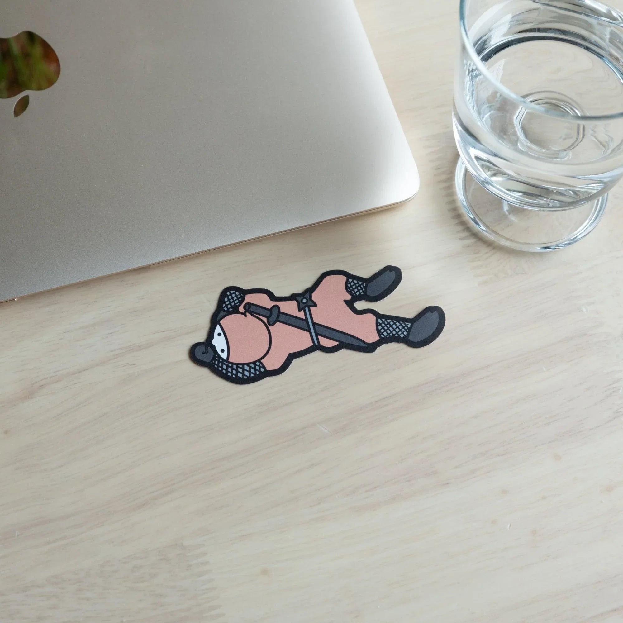 Pink ninja die-cut card lying flat on a wood desk in crawling pose, with a closed MacBook and water glass in background