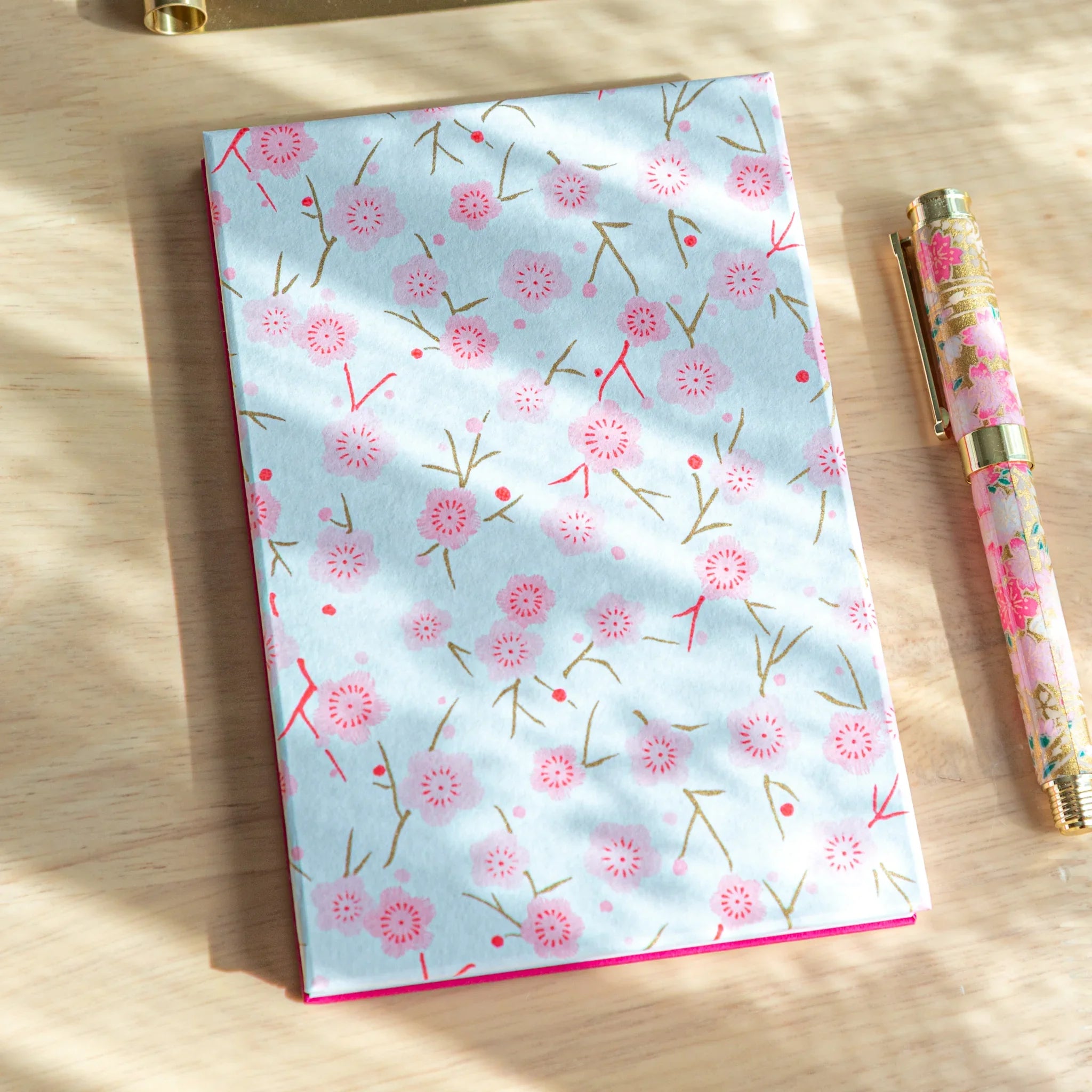 Pink Plum Blossoms washi goshuincho front cover in light blue with pink plum and gold branch print on light wood surface