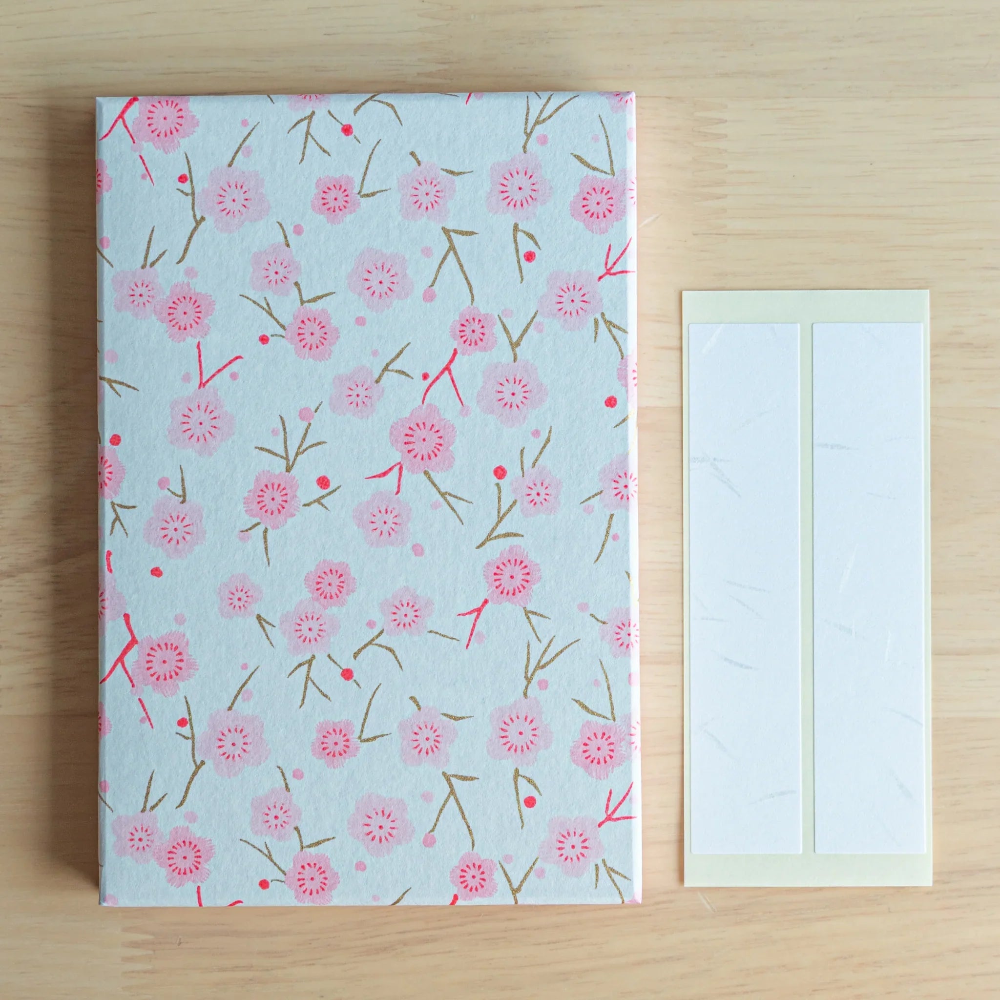 Pink Plum Blossoms washi goshuincho front cover flat lay on wood surface beside two included cream washi name label strips