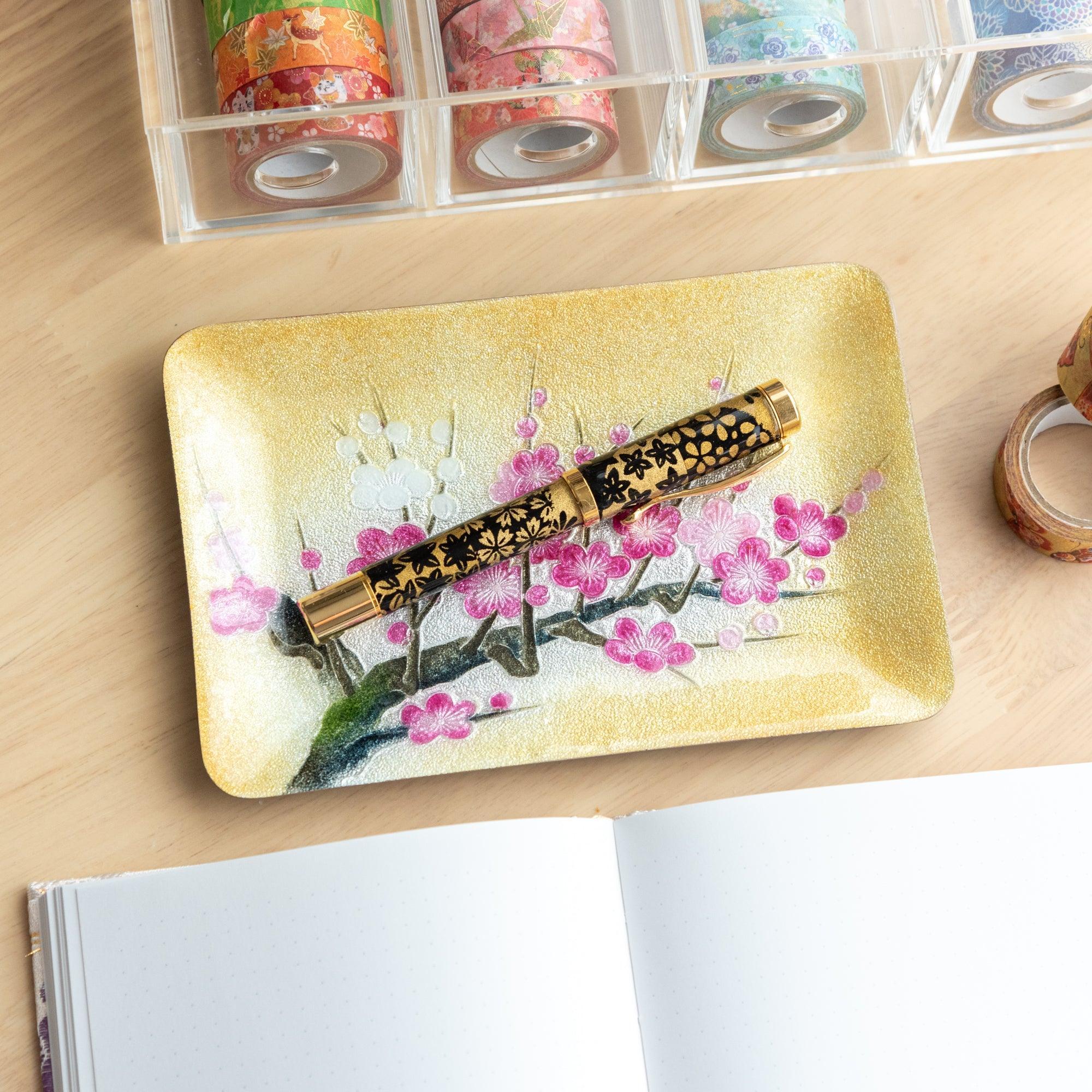 A black and gold fountain pen resting on a tray adorned with vibrant pink plum blossoms, showcasing elegance in Japanese design.