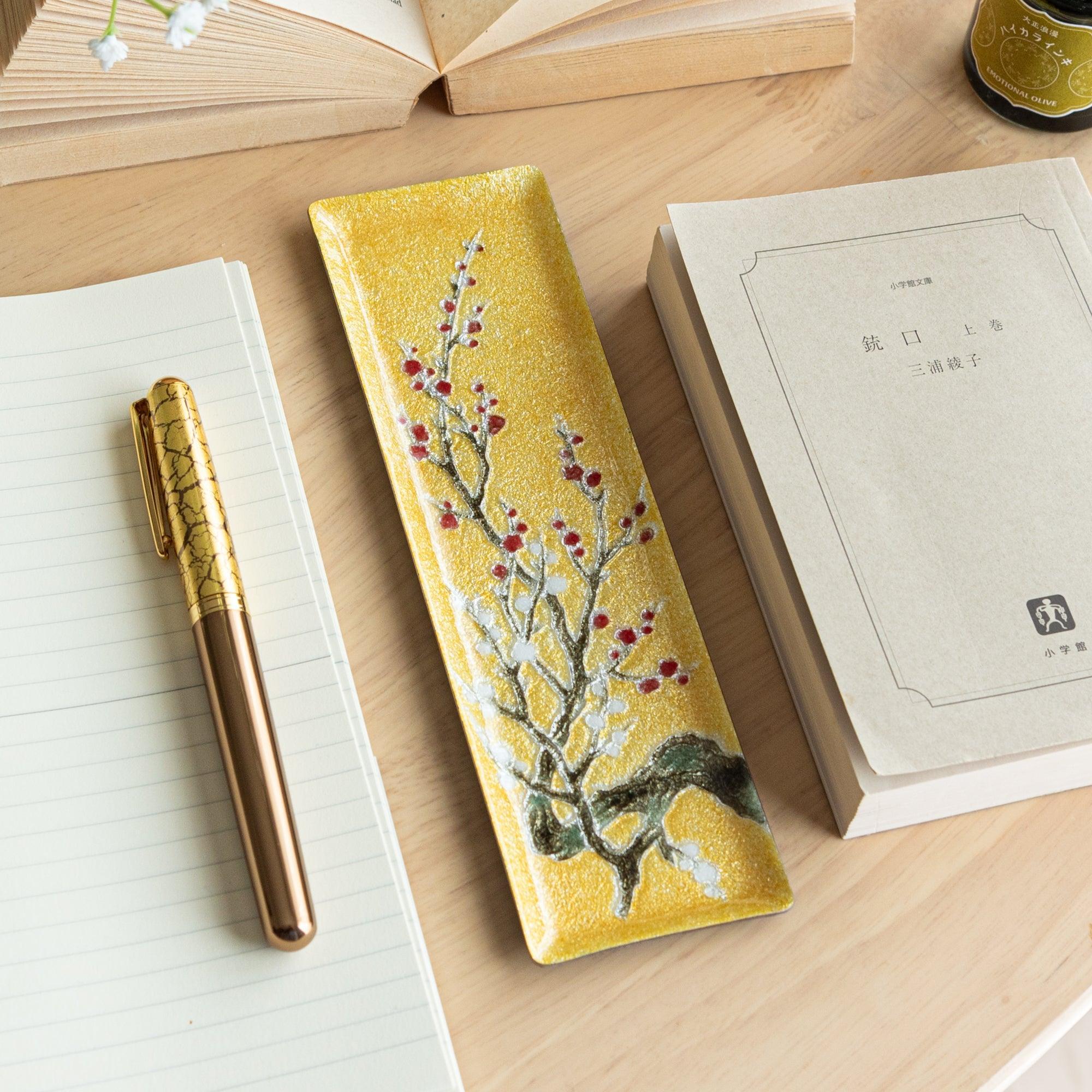 Japanese shippo ware slim pen tray with white and red plum blossom design on gold enamel, on wood desk with notebook and book