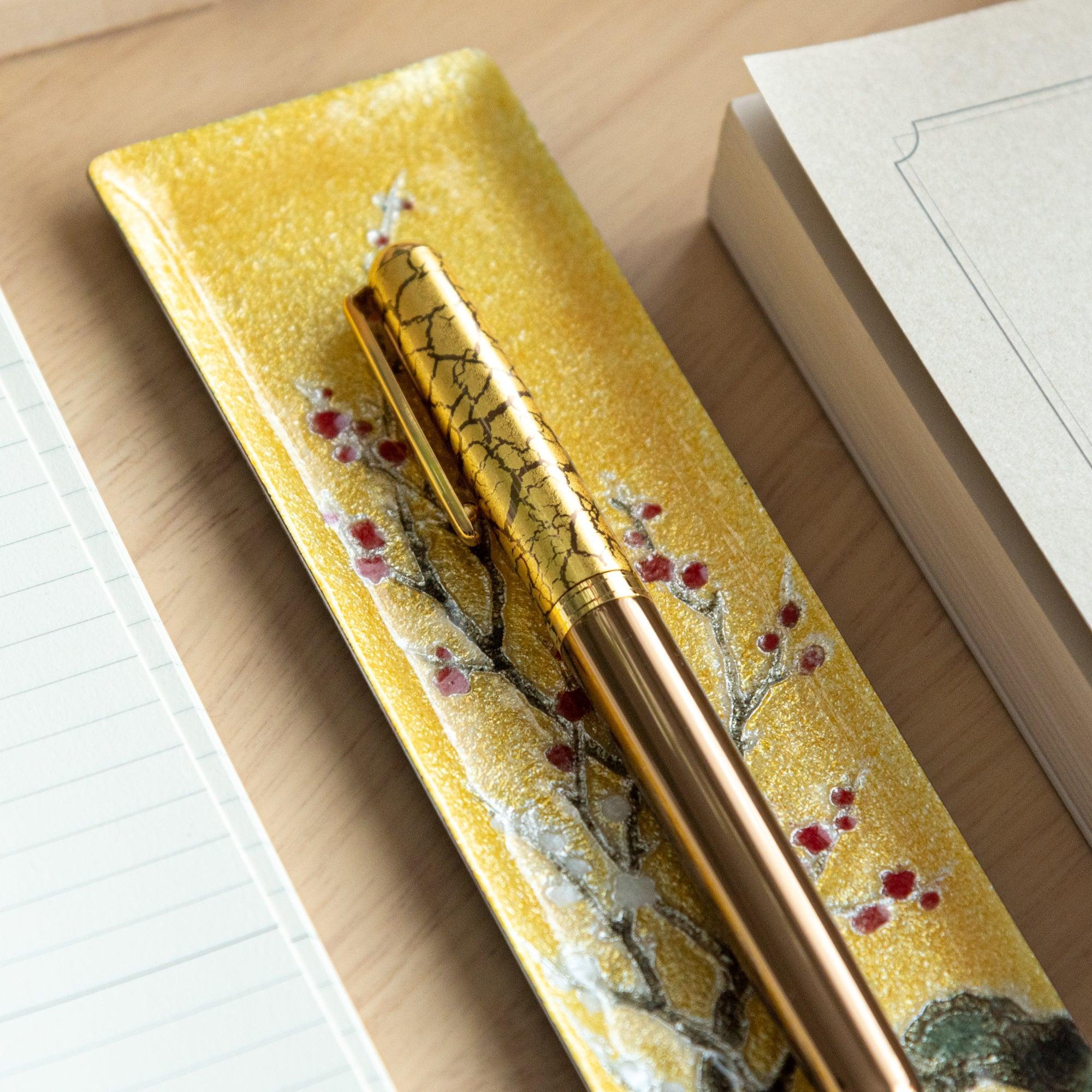 Close-up of gold enamel slim pen tray with red plum branch design, gold crackle pen on tray, notebooks flanking on wood desk