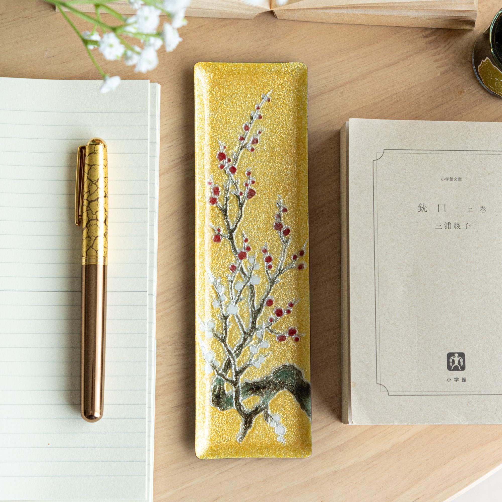 Gold enamel slim pen tray with plum branch and red berry design, overhead flat lay, gold fountain pen on notebook to left
