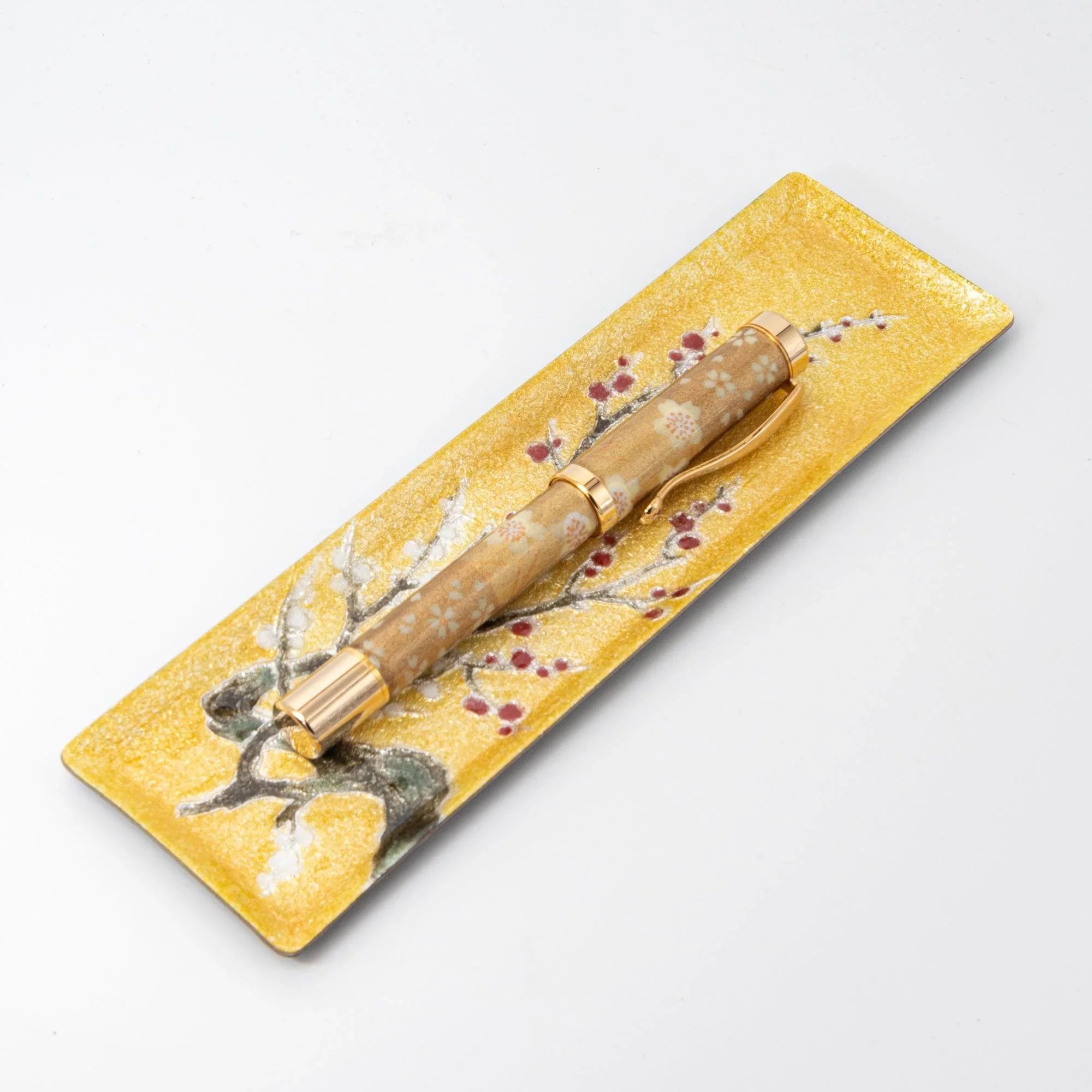 Gold enamel slim pen tray with plum branch design on white background, angled view, floral pen resting on tray