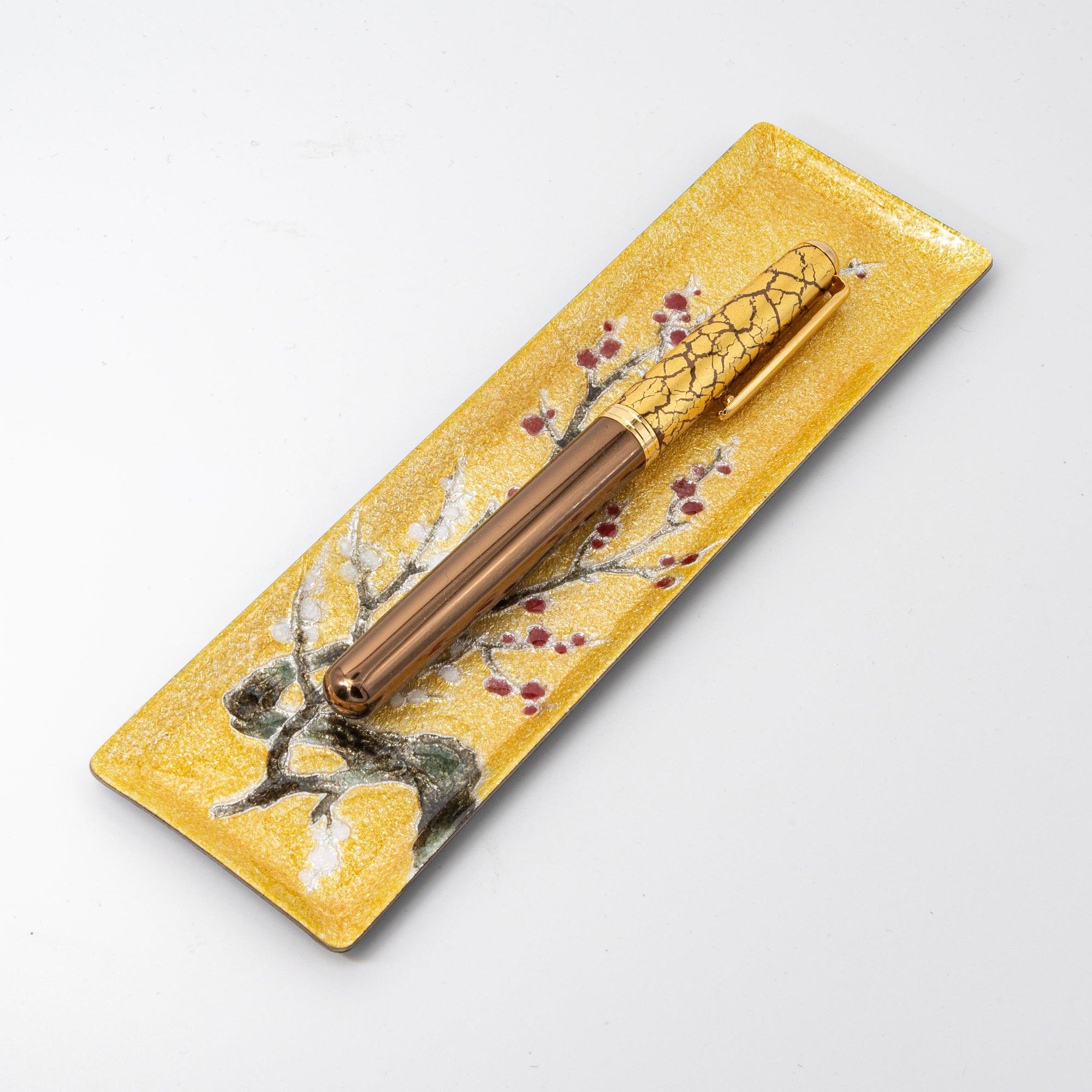 Gold enamel slim pen tray with plum branch design on white background, angled view, gold crackle fountain pen resting on tray