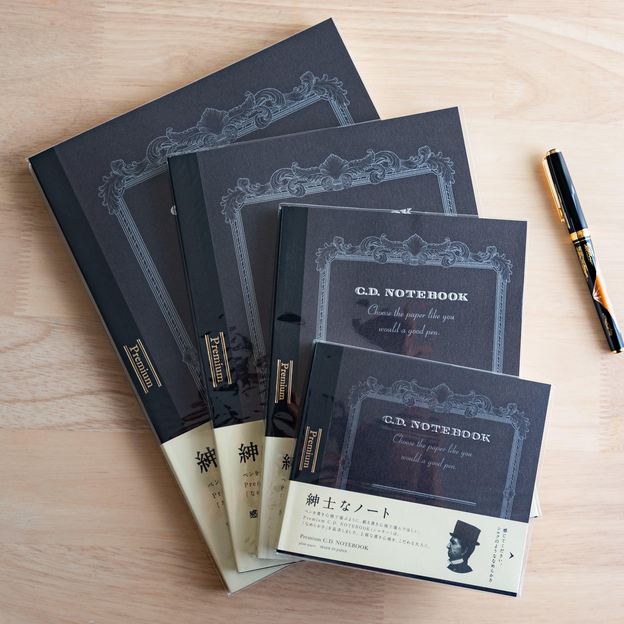 Four black CD notebooks in various sizes are arranged on a wooden surface beside a black and gold fountain pen