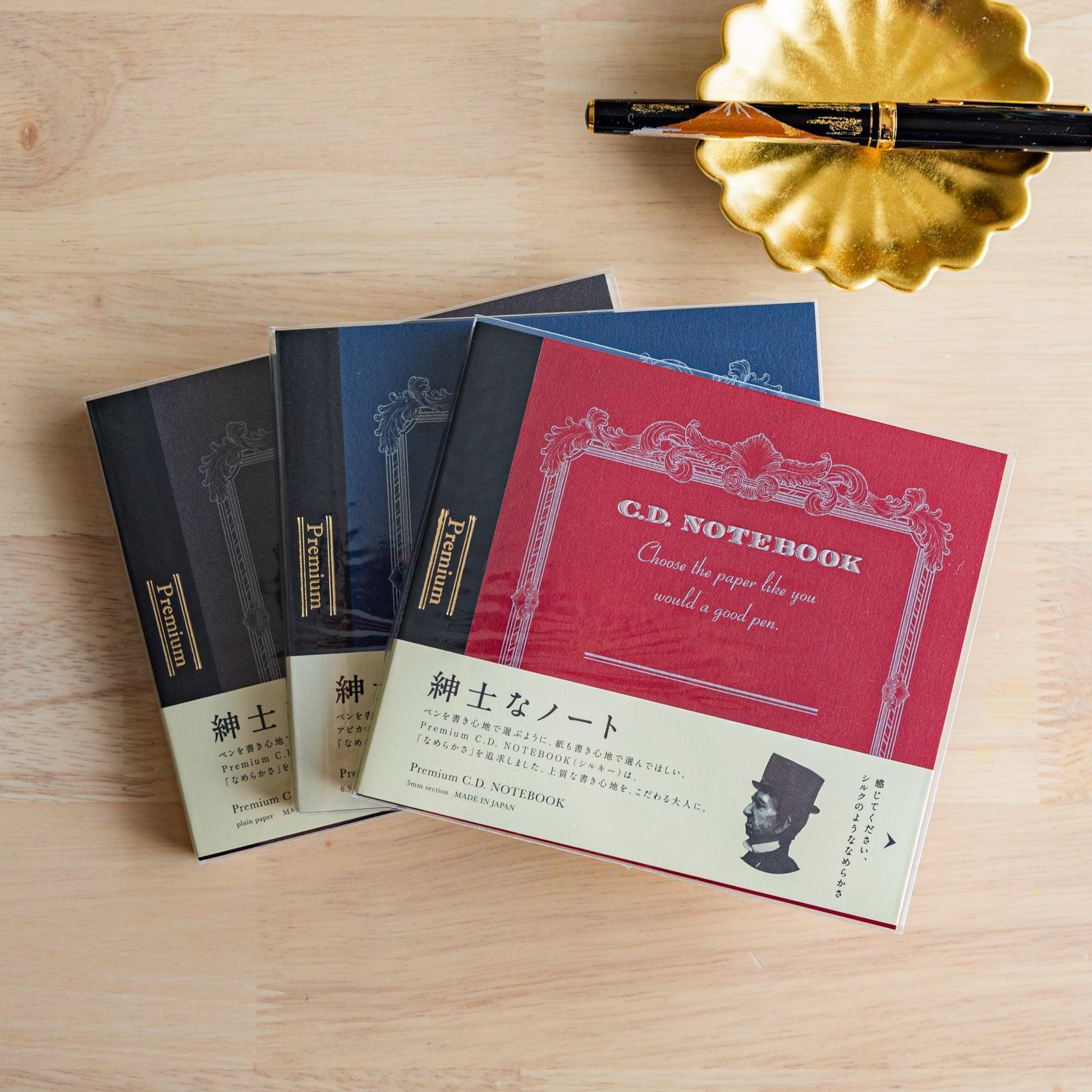 Three Premium CD Japanese notebooks in black, red, and blue covers with a fountain pen, showcasing refined Japanese stationery