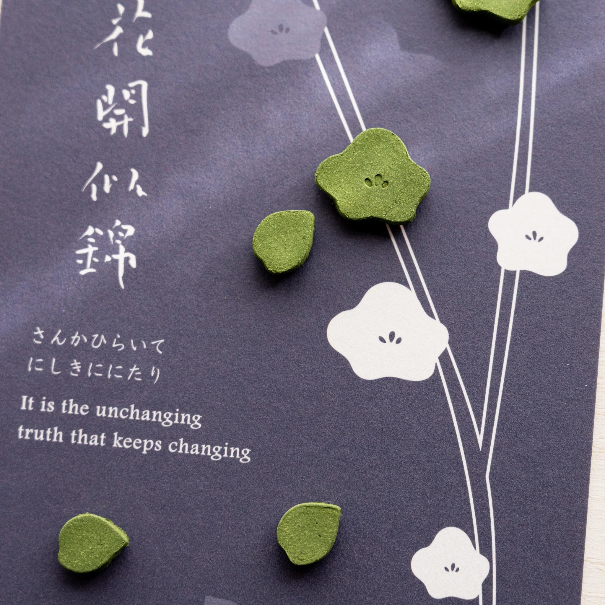 Close-up of plum variant pressed matcha incense card with white calligraphy on dark navy paper and small plum-shape pieces