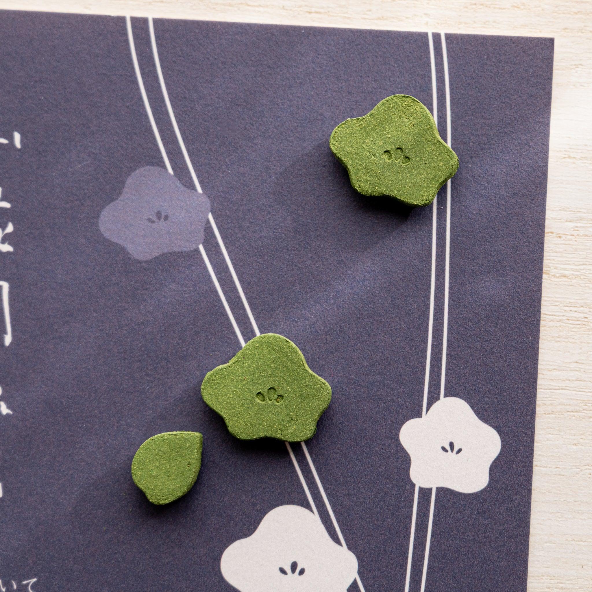 Close-up of three small green pressed matcha incense pieces in plum flower and leaf shapes on dark navy card