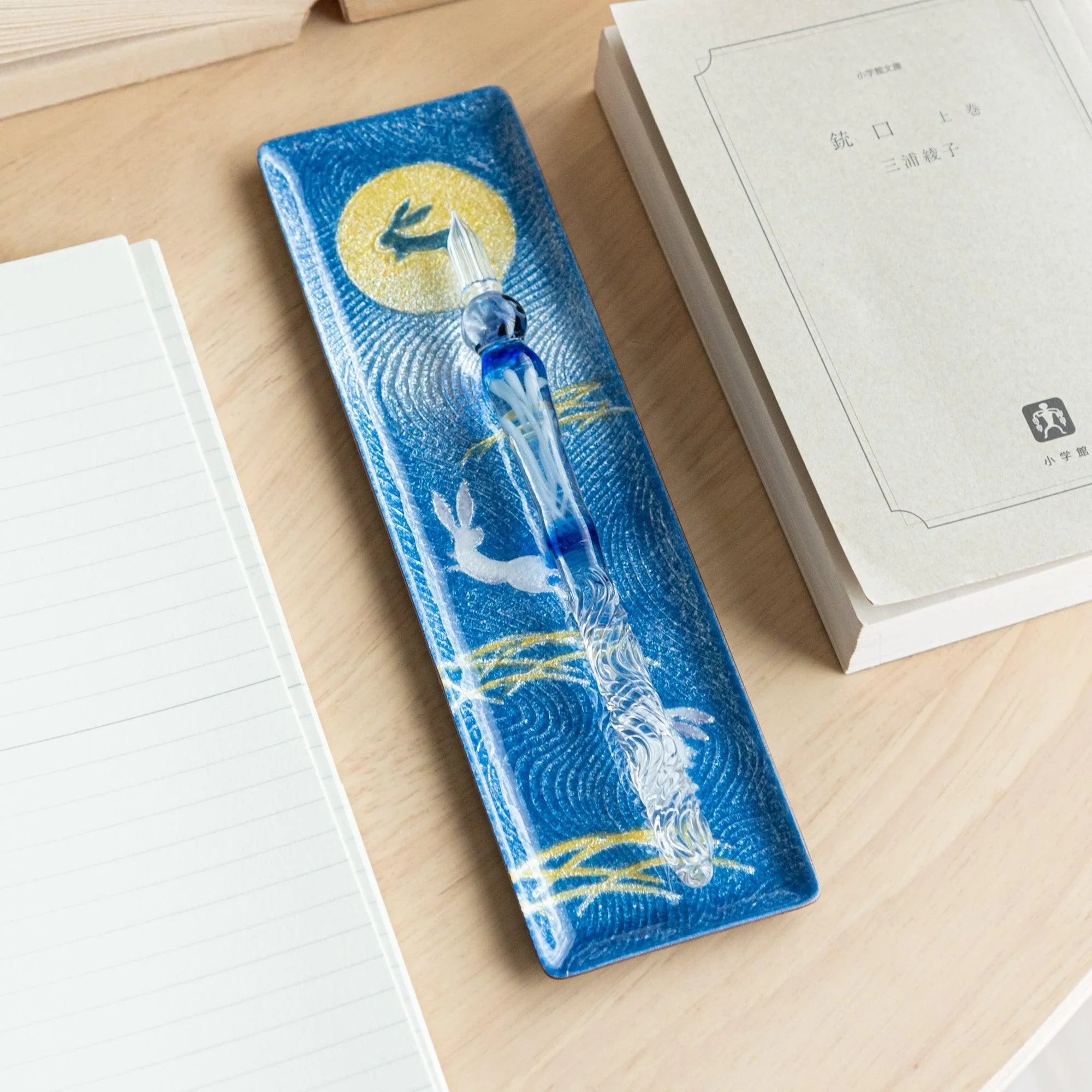 Dark blue enamel slim pen tray with rabbit and moon design, blue glass pen on tray, wood desk with notebook and Japanese book