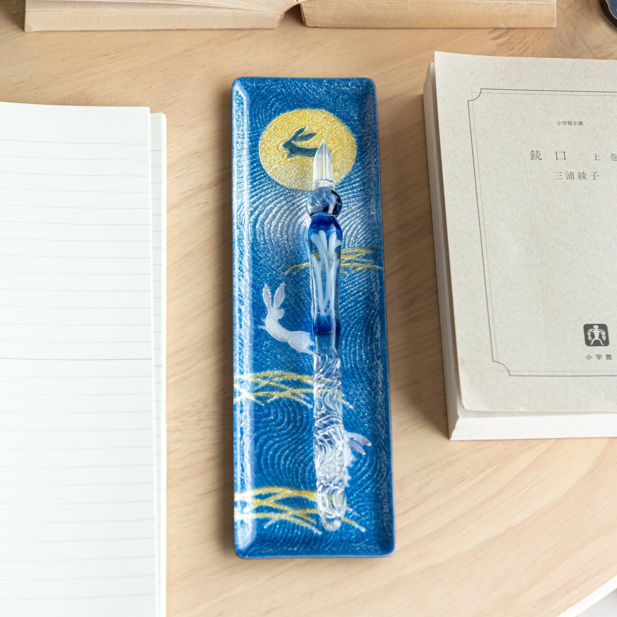 A top-down view of a blue Japanese stationery tray with a moon and rabbit design, showcasing golden grass and a glass dip pen.