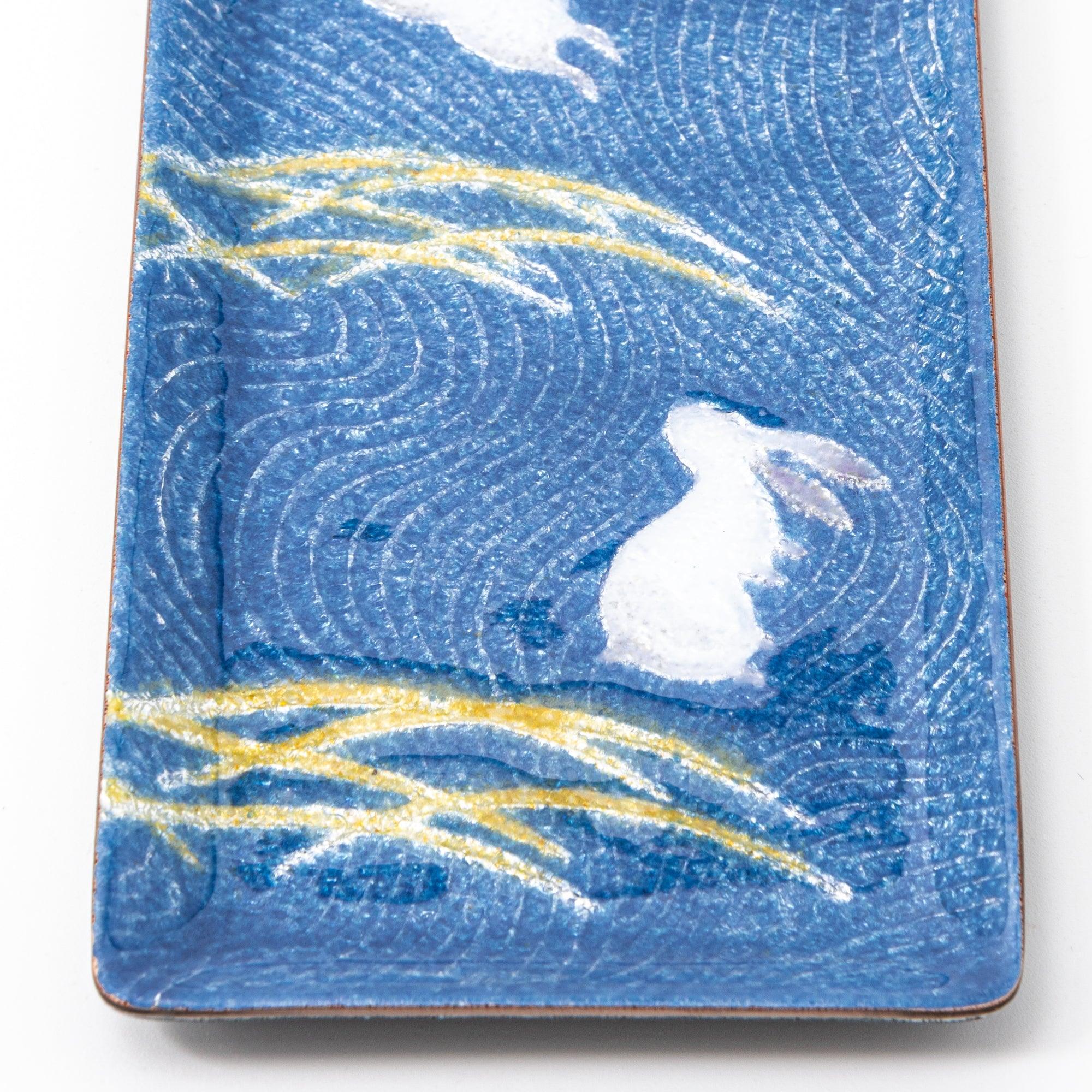 Close-up of dark blue enamel tray showing sitting white rabbit and gold pampas grass on blue wave pattern