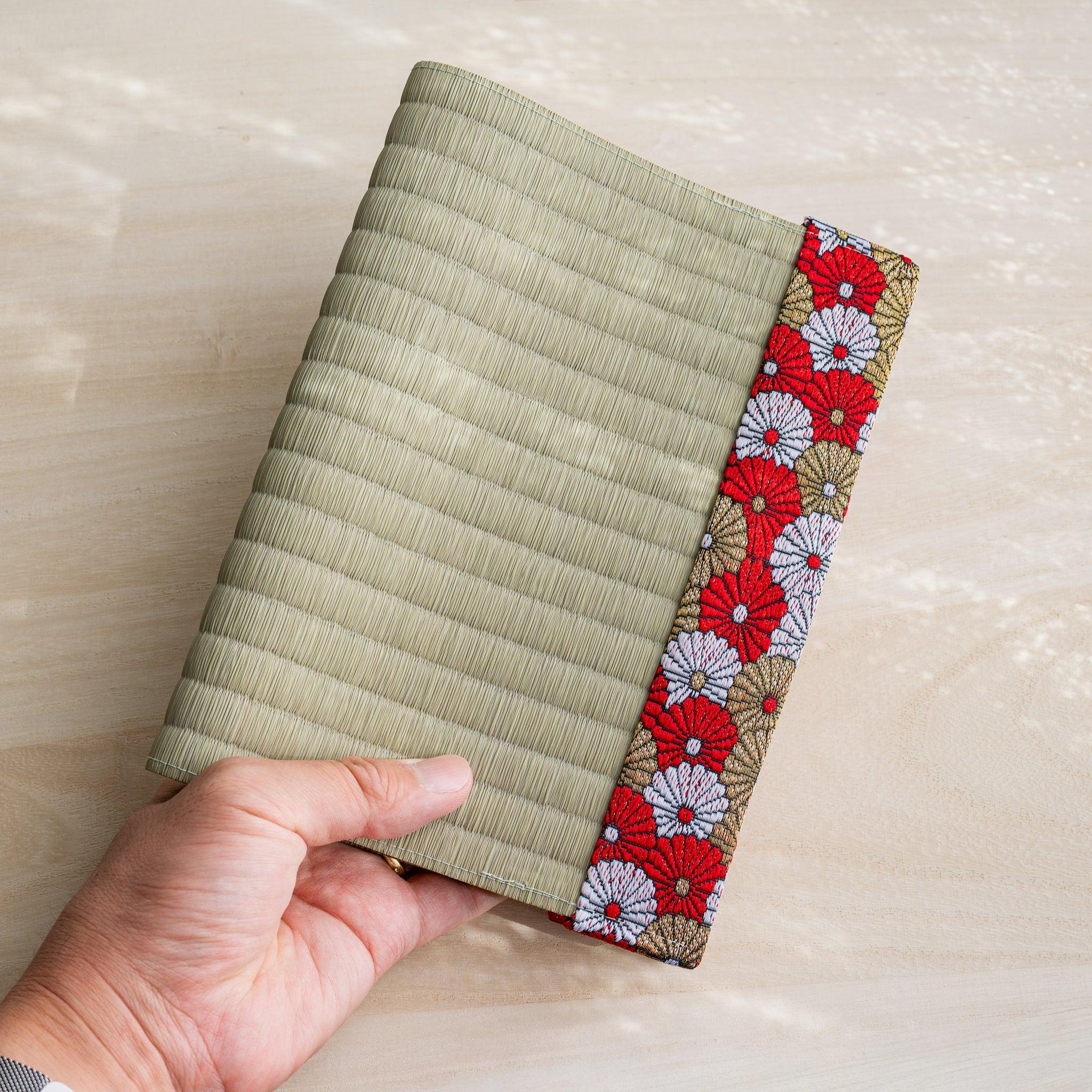 Hand holding closed tatami book cover, green rush straw front with red white and gold Japanese chrysanthemum band