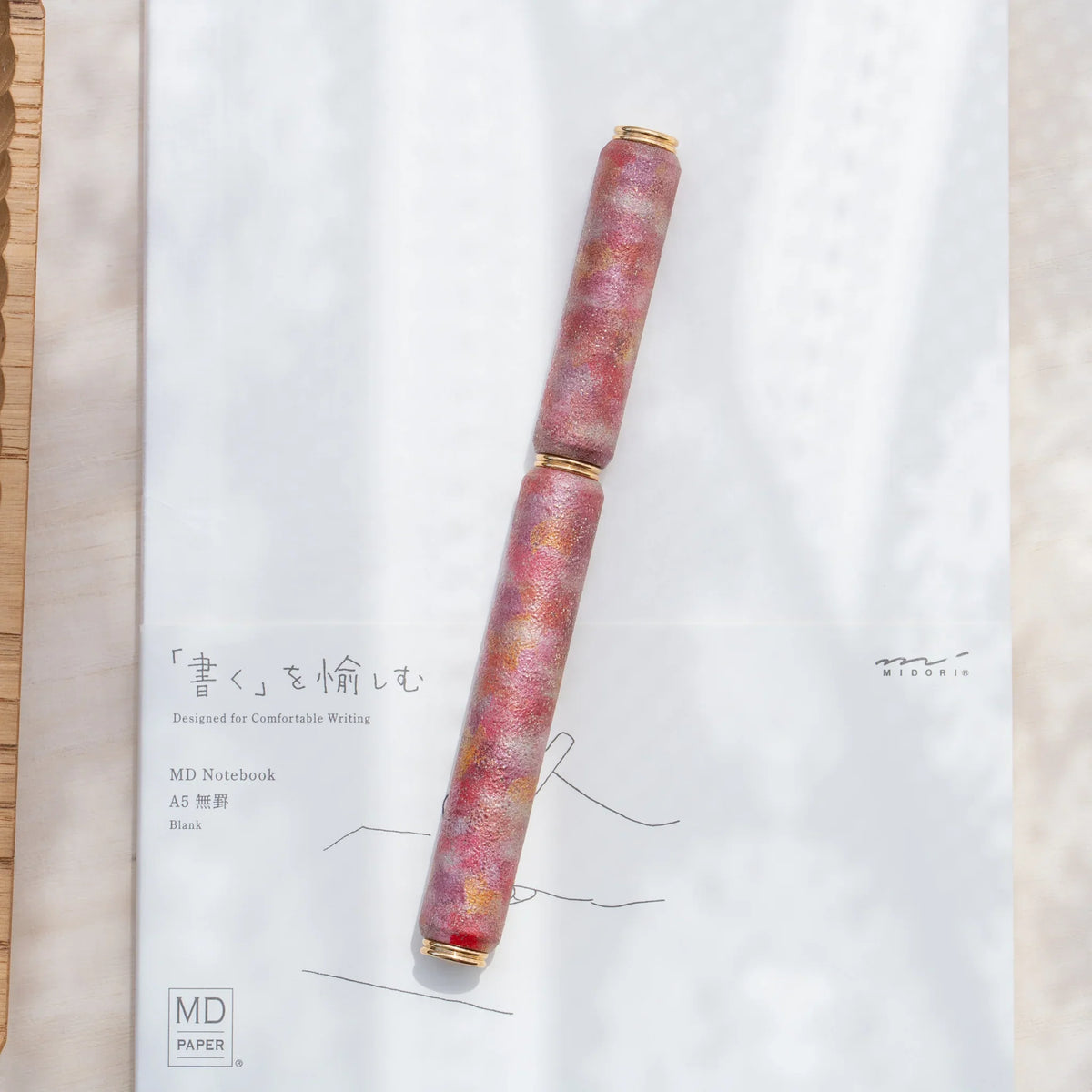 Red marbled urushi lacquered rollerball pen in zelkova wood, resting on a Japanese notebook