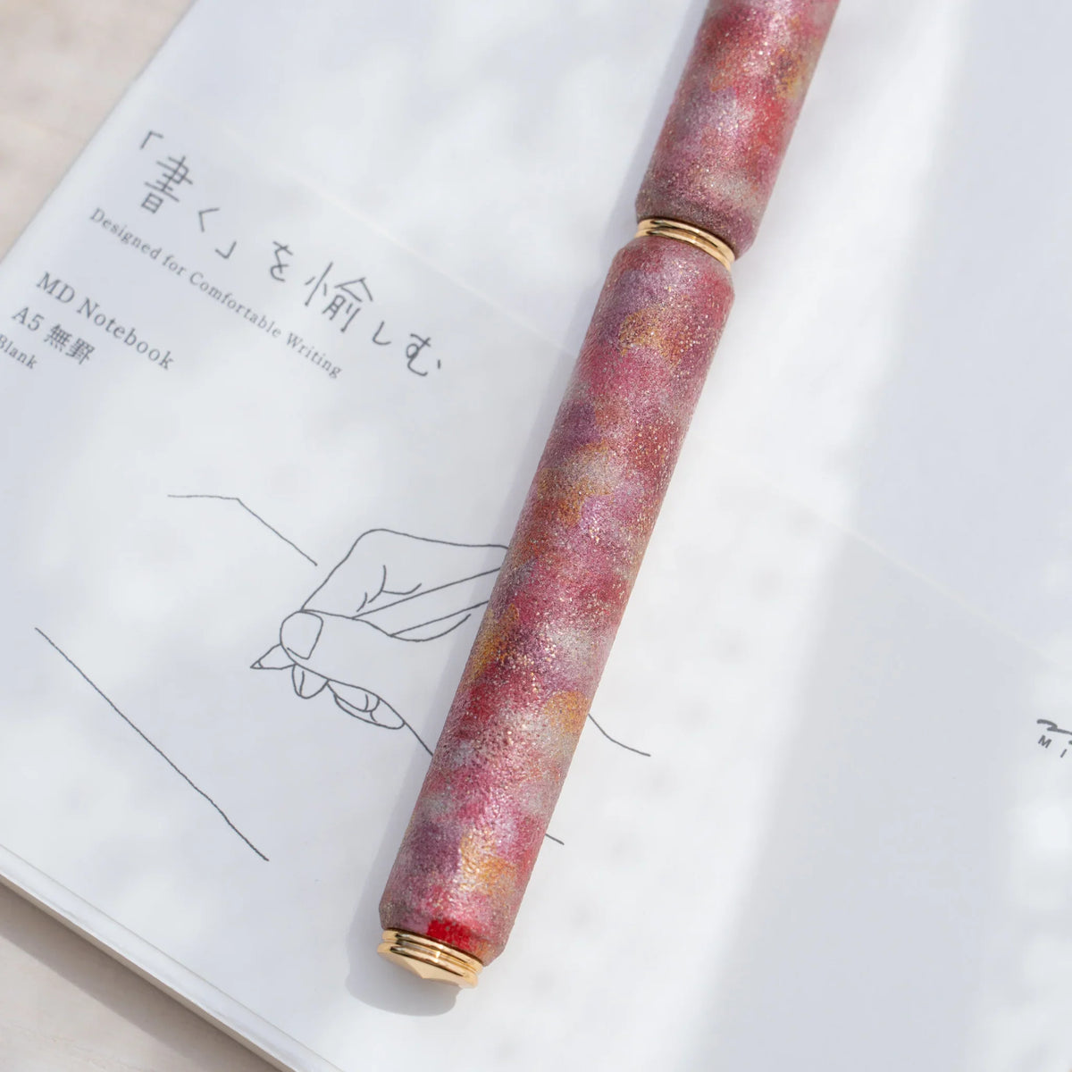 Red marbled urushi lacquered rollerball pen in zelkova wood, resting on a Japanese notebook