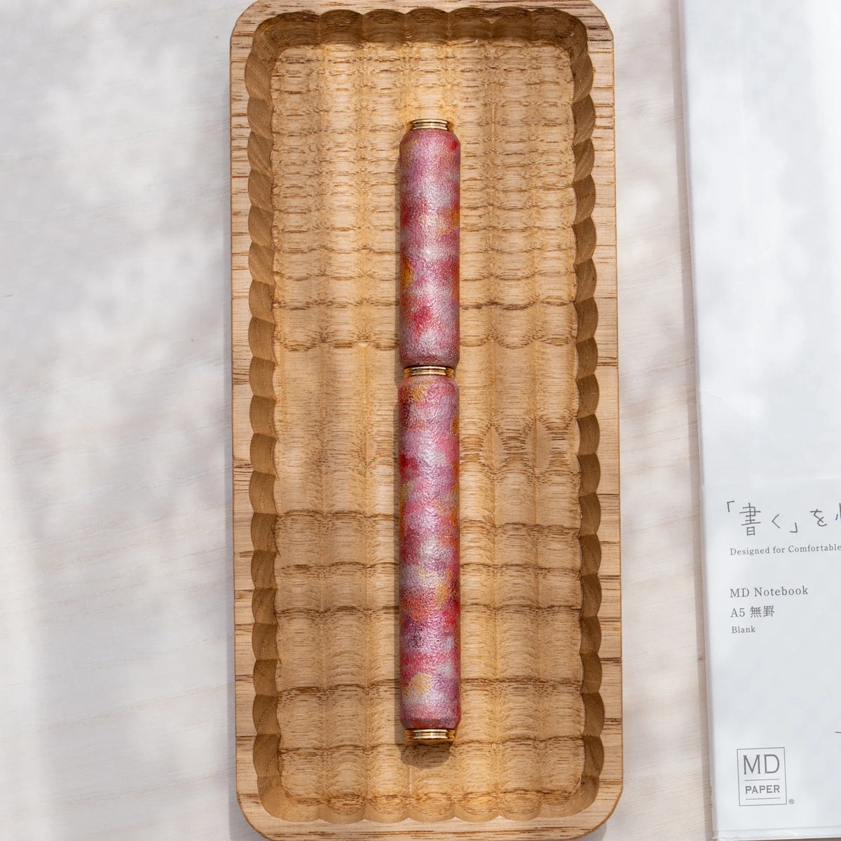 Red marbled urushi lacquered rollerball pen in zelkova wood, shown in a carved wooden tray with a Japanese notebook