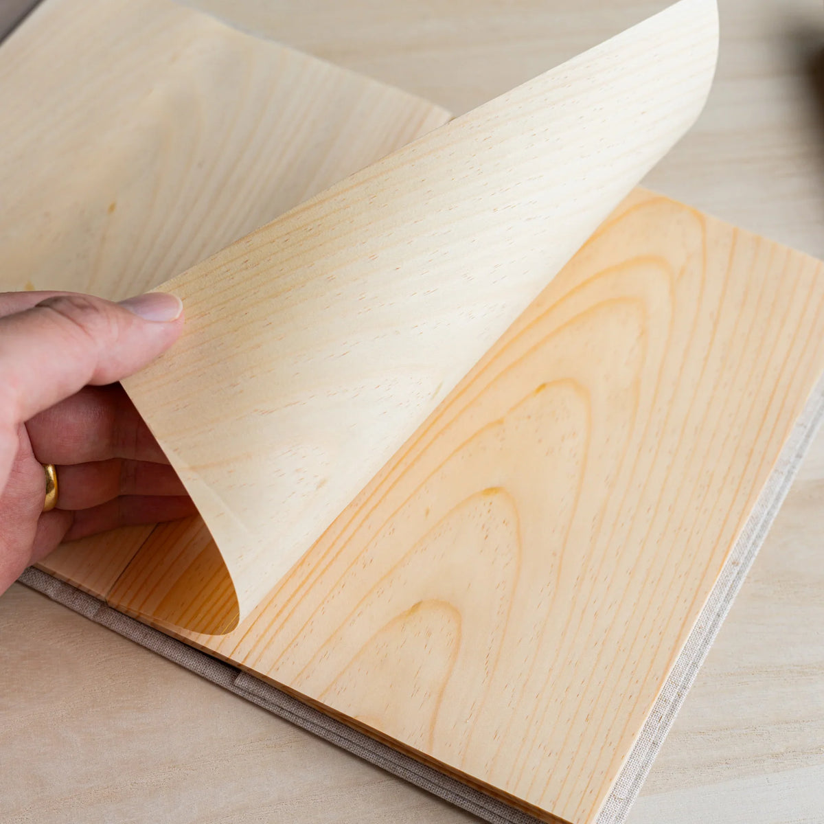 Hand lifting a thin wood sheet page of the Red Pine Wooden Page Sketchbook, showing grain on the page below