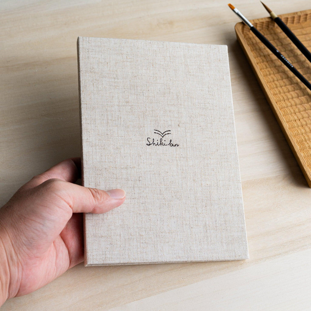 Hand holding the Red Pine Wooden Page Sketchbook Natural, linen hardcover with Shiki bun logo