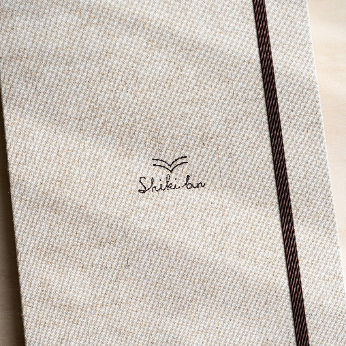 Close-up of the Natural cover, woven linen texture with printed Shiki bun logo and dark brown elastic band