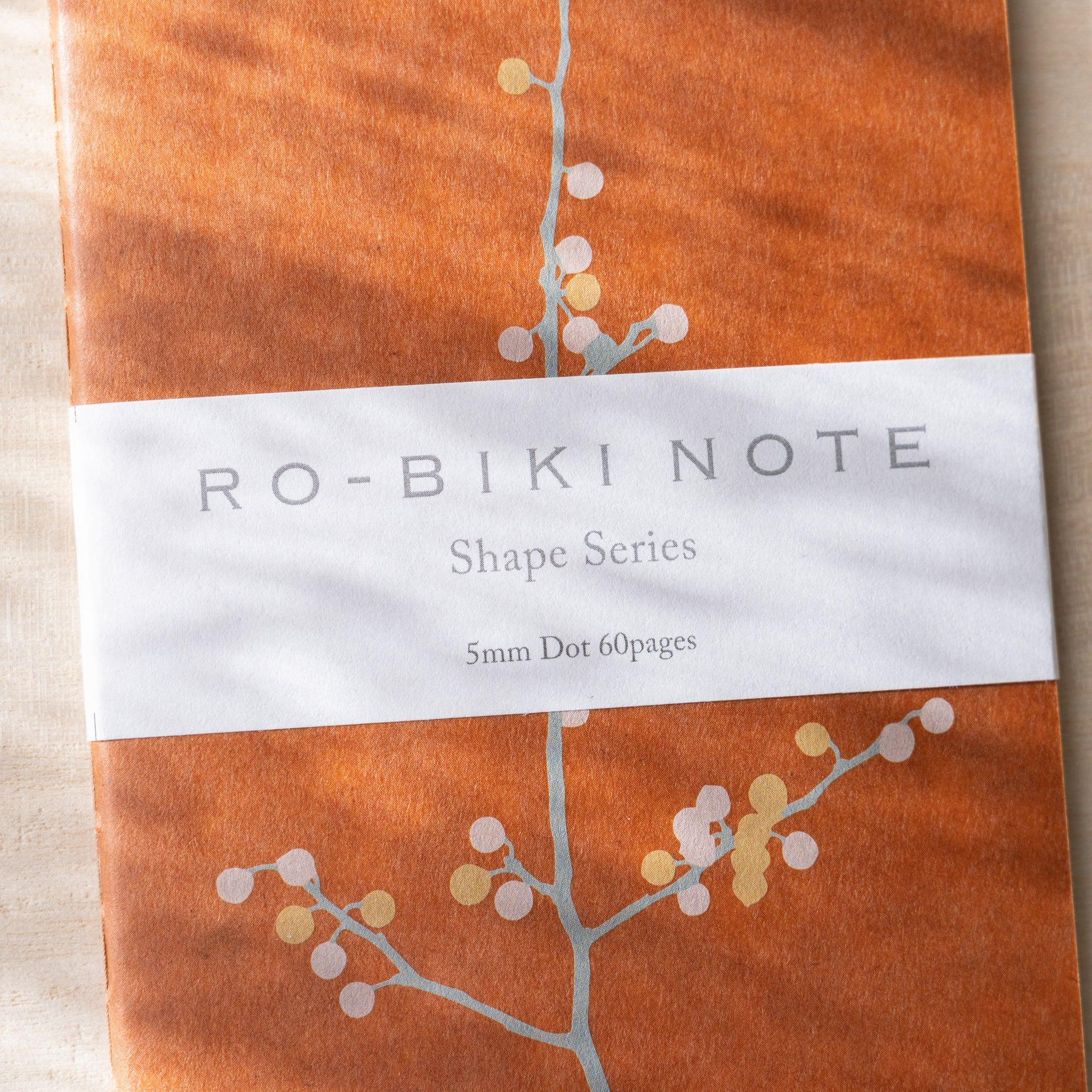 RO-BIKI NOTE Japanese notebook close-up, terracotta waxed cover with branch and berry dots, belly band with Shape Series text