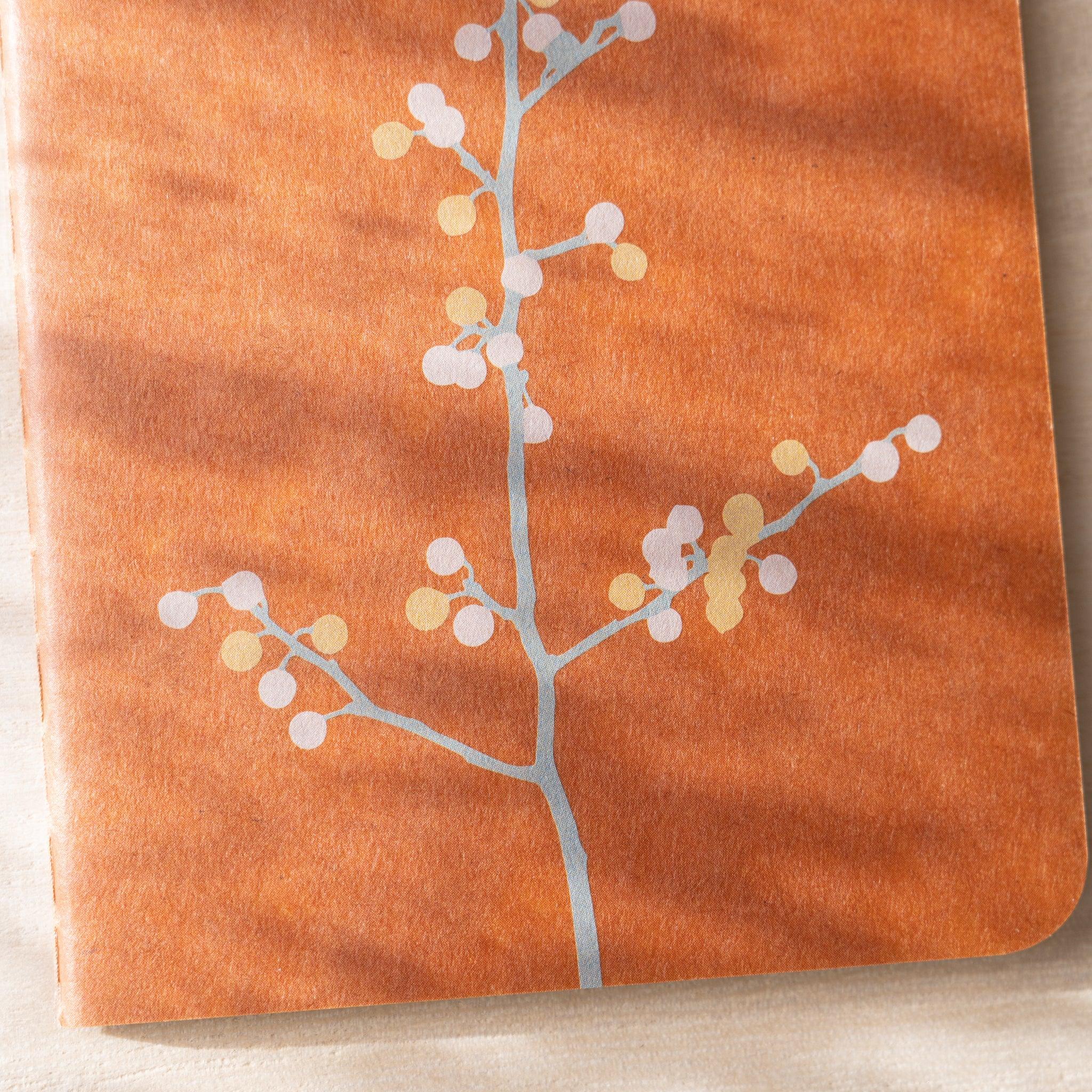 Close-up of RO-BIKI NOTE cover showing branch with silver and gold botanical detail, waxed finish visible