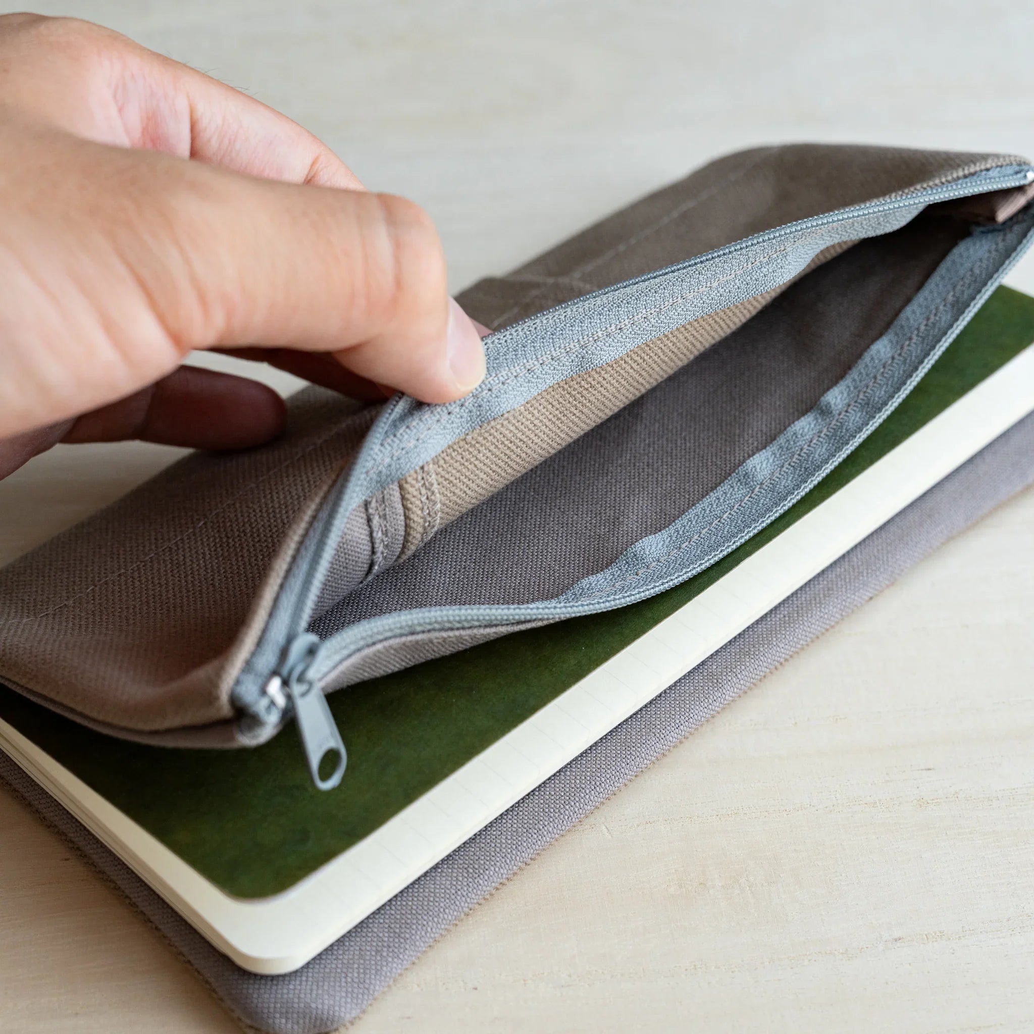 Hand opening the pale blue side zip pocket of the RO-BIKI clay beige canvas cover over a green notebook