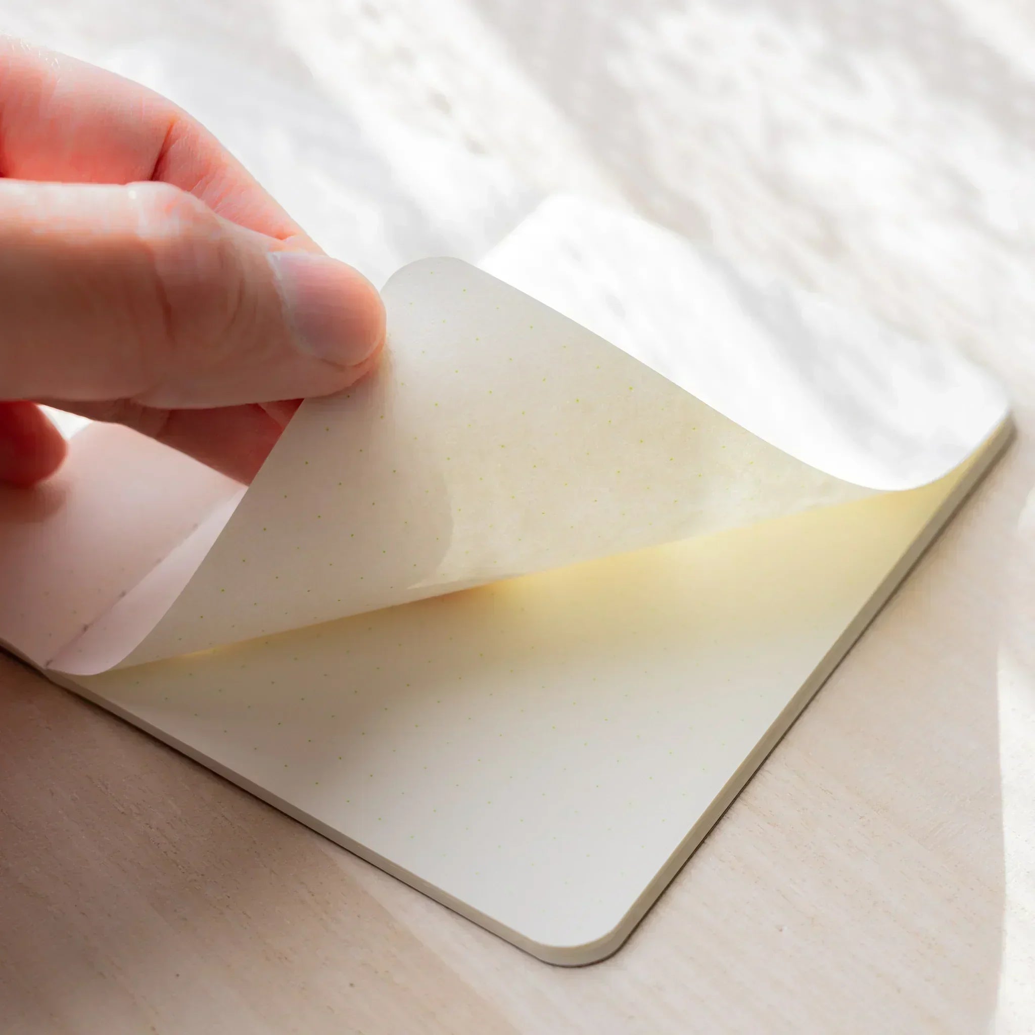 Hand peeling back the corner of an ivory page in the RO-BIKI NOTE, showing paper thickness and rounded corners