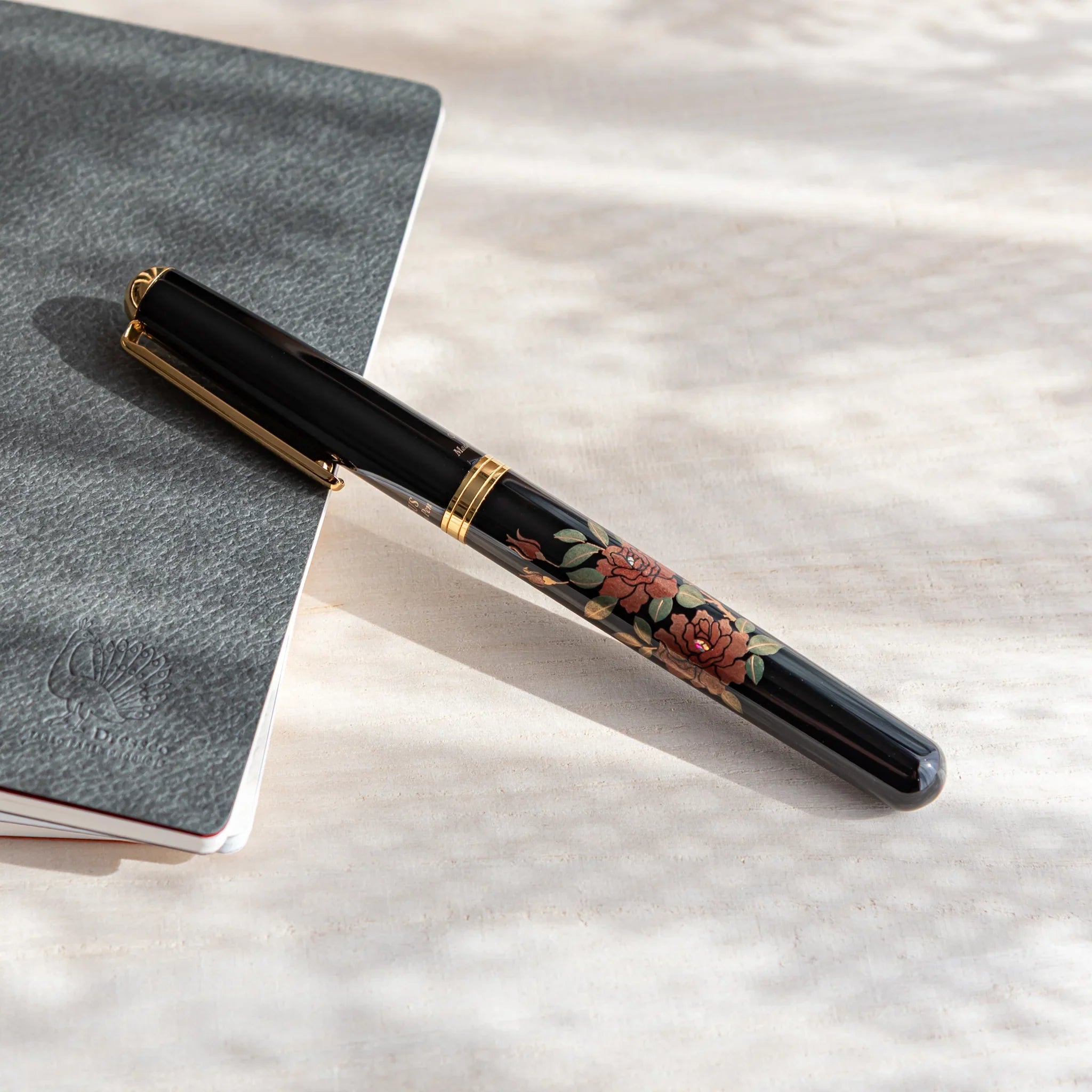 Black Japanese fountain pen with rose floral lacquer design resting diagonally on a notebook under soft light.